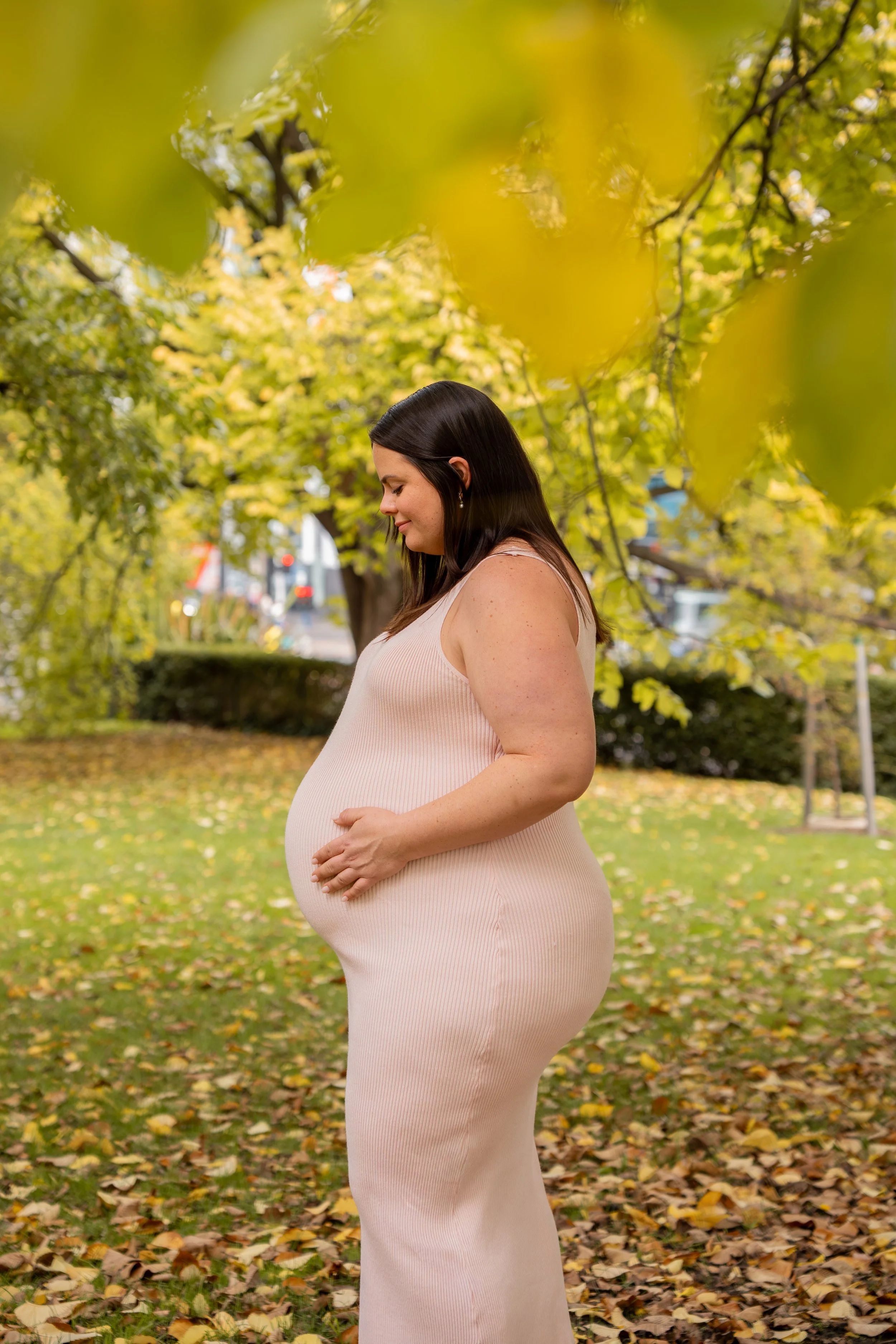 Pregnant woman in a light pink dress standing in park with yellow and green leaves on trees and ground.