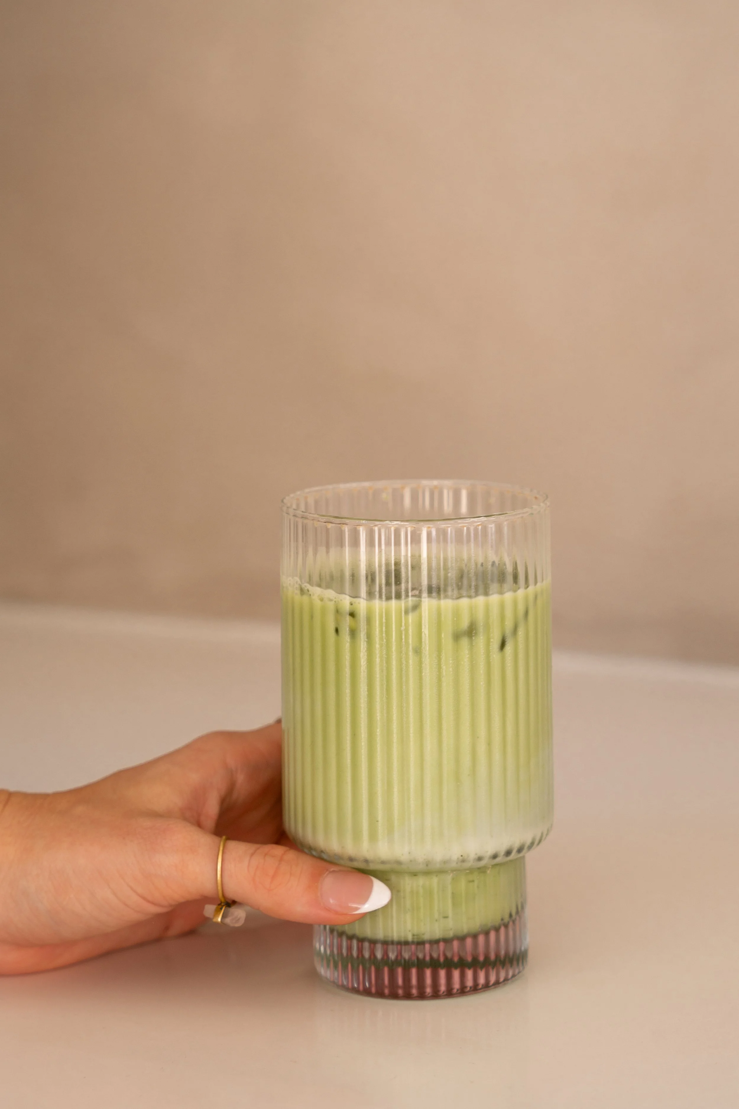 A hand holding a ribbed glass containing a green beverage, with two other glasses stacked below, all with the same pattern, against a simple beige background.