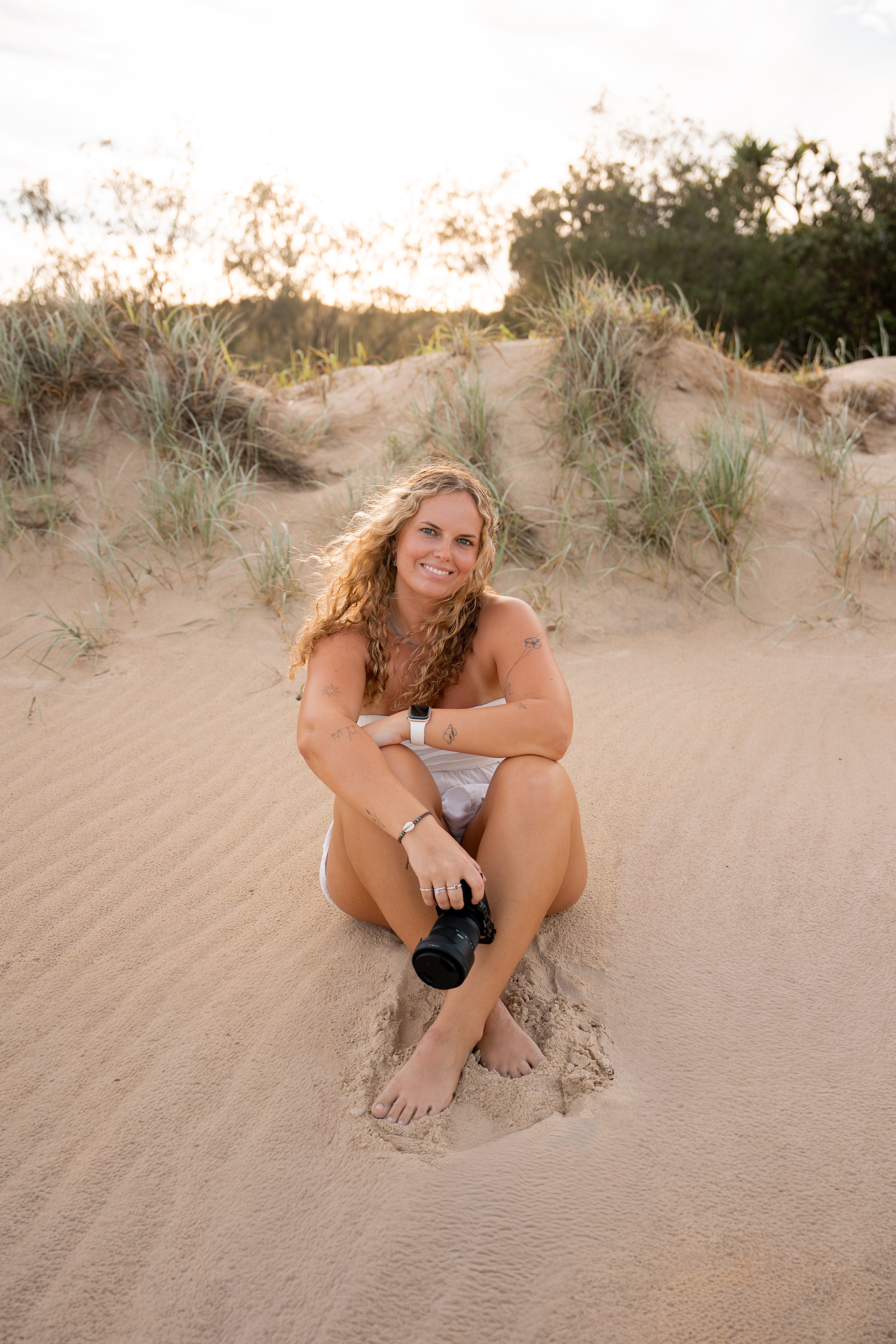 A woman with curly blonde hair sitting on sandy beach dunes, smiling and holding a camera, with a backdrop of sand dunes and trees during sunset.