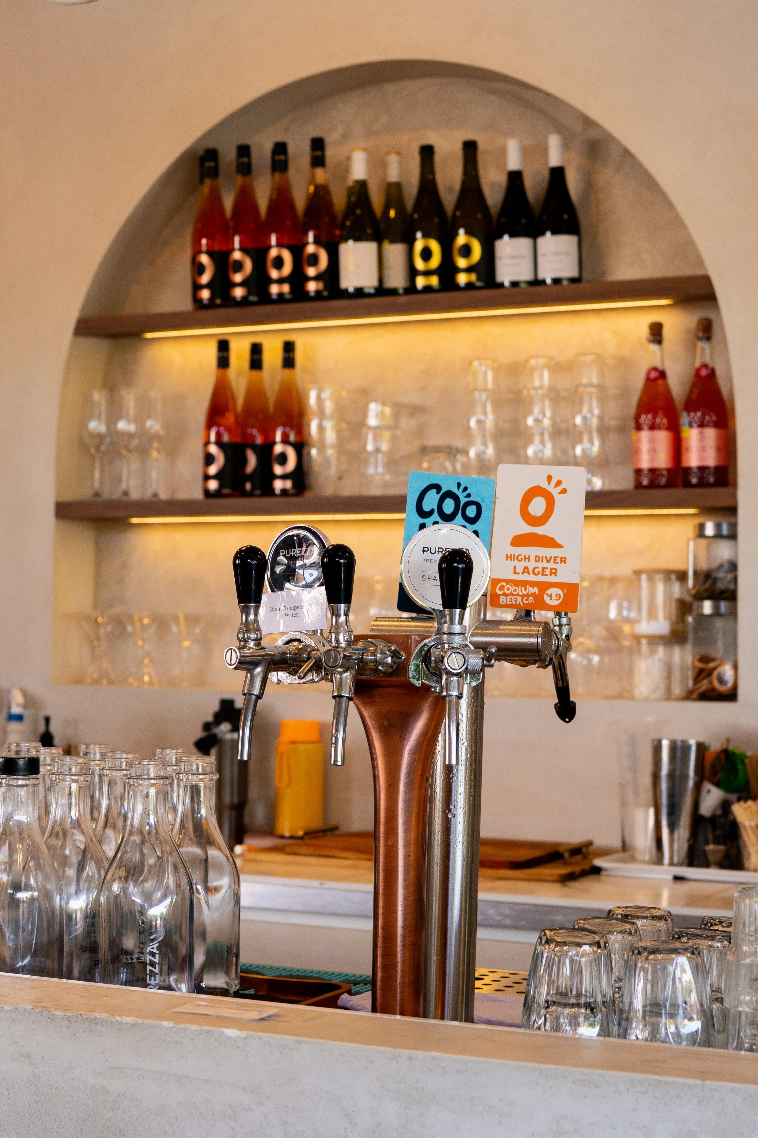 Bar counter with glass bottles, bar taps, and glasses. Bottles on shelves in the background and bar tools on the counter.