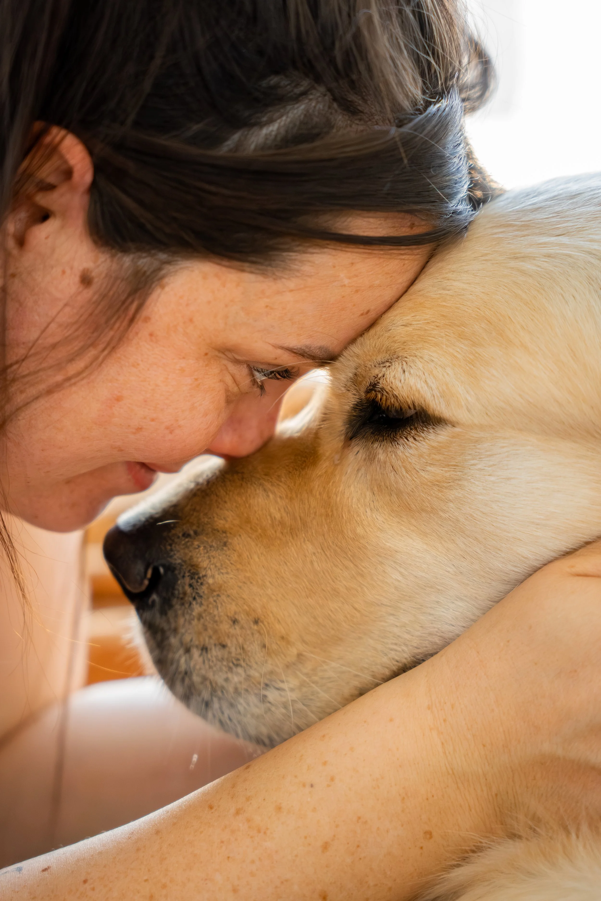 A woman with dark brown hair and freckles touching foreheads with a large, light-colored dog, both with eyes closed, creating an intimate and tender moment.