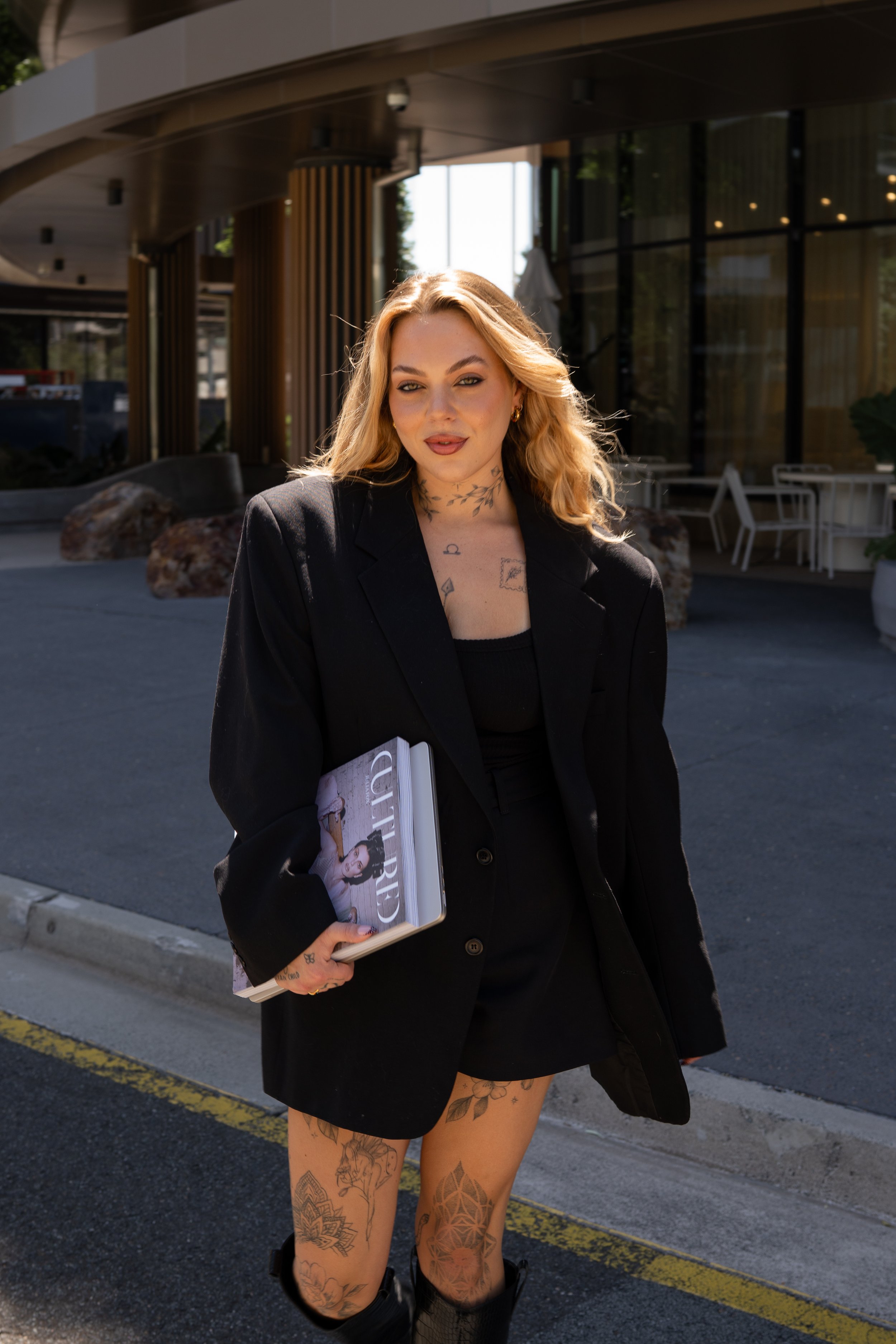 A woman with blonde hair and tattoos on her legs and chest, wearing a black blazer and holding a Vogue magazine, standing outside a modern building with glass windows and wooden pillars.
