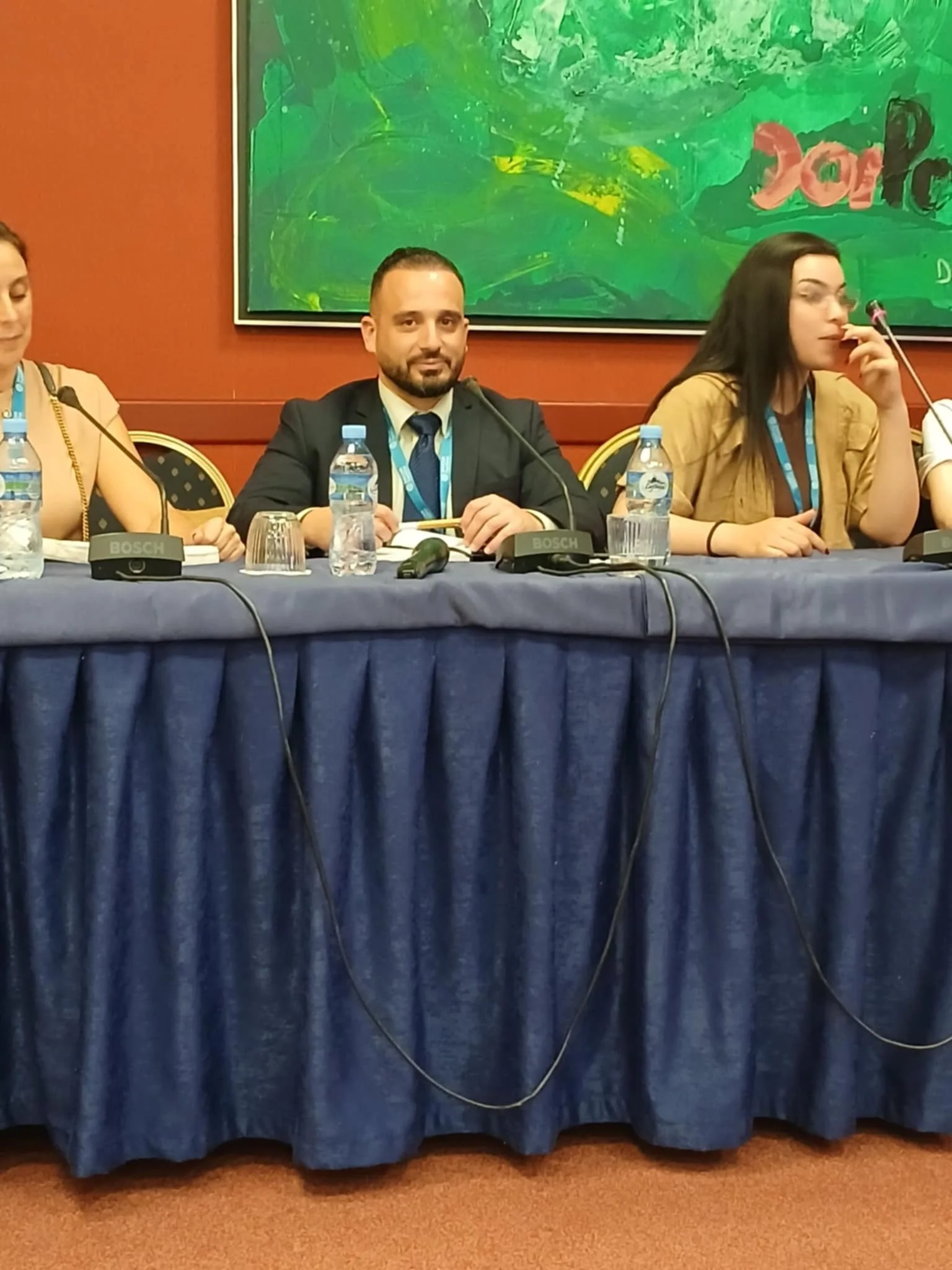 A panel of three people sitting at a table during a conference or meeting. The man in the center is wearing a suit and tie, with a water bottle and a microphone in front of him. To his right is a woman with dark hair, glasses, and a beige jacket, also with a water bottle. The table is covered with a dark blue cloth, and there are microphones with the brand Bosch placed in front of each person. Behind them, a large green abstract painting and a maroon wall are visible.