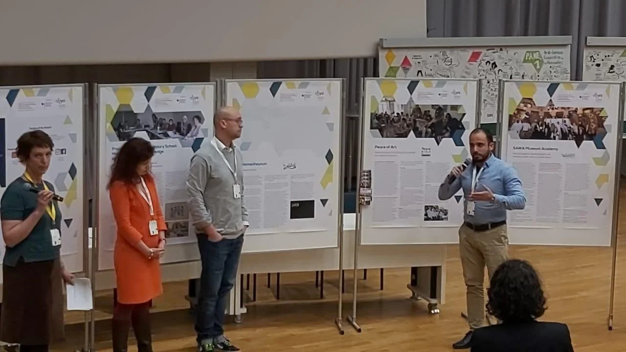 People giving a presentation at an academic conference or exhibit with poster boards in the background.