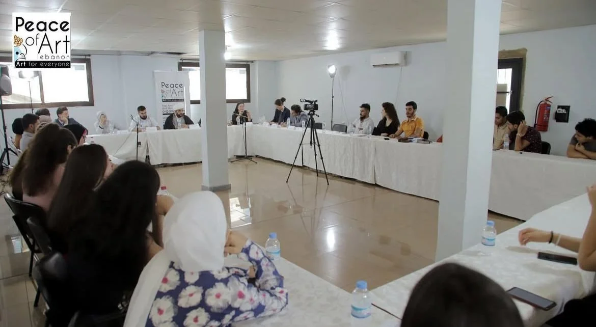 A group of people sitting around a U-shaped conference table during a meeting or discussion. There is a camera set up in the center of the room, and a banner on the wall that reads 'Peace of Art Lebanon'.