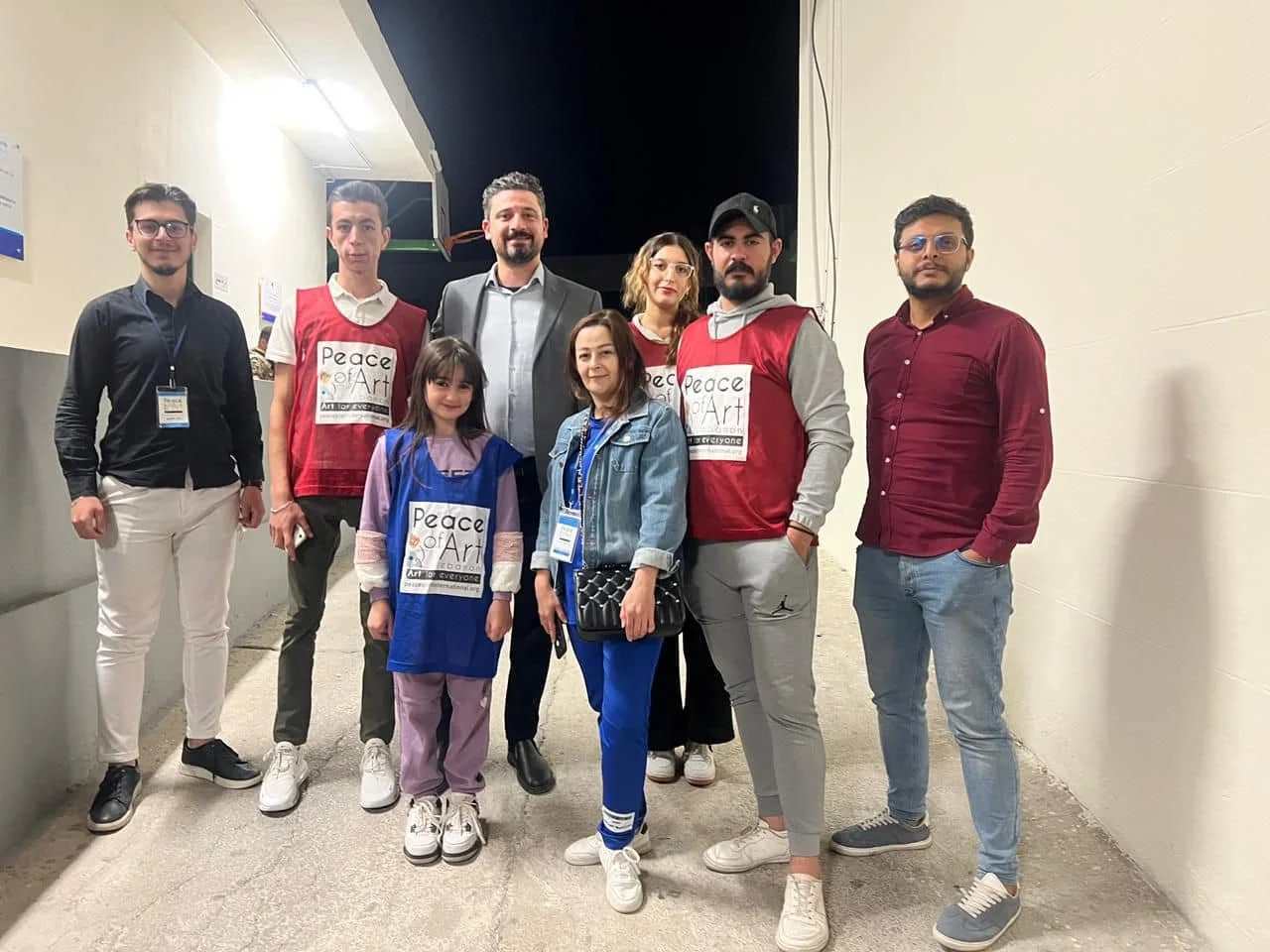 Group of nine people standing together in a hallway at an event, some wearing 'Peace of Art' vests, smiling at the camera.