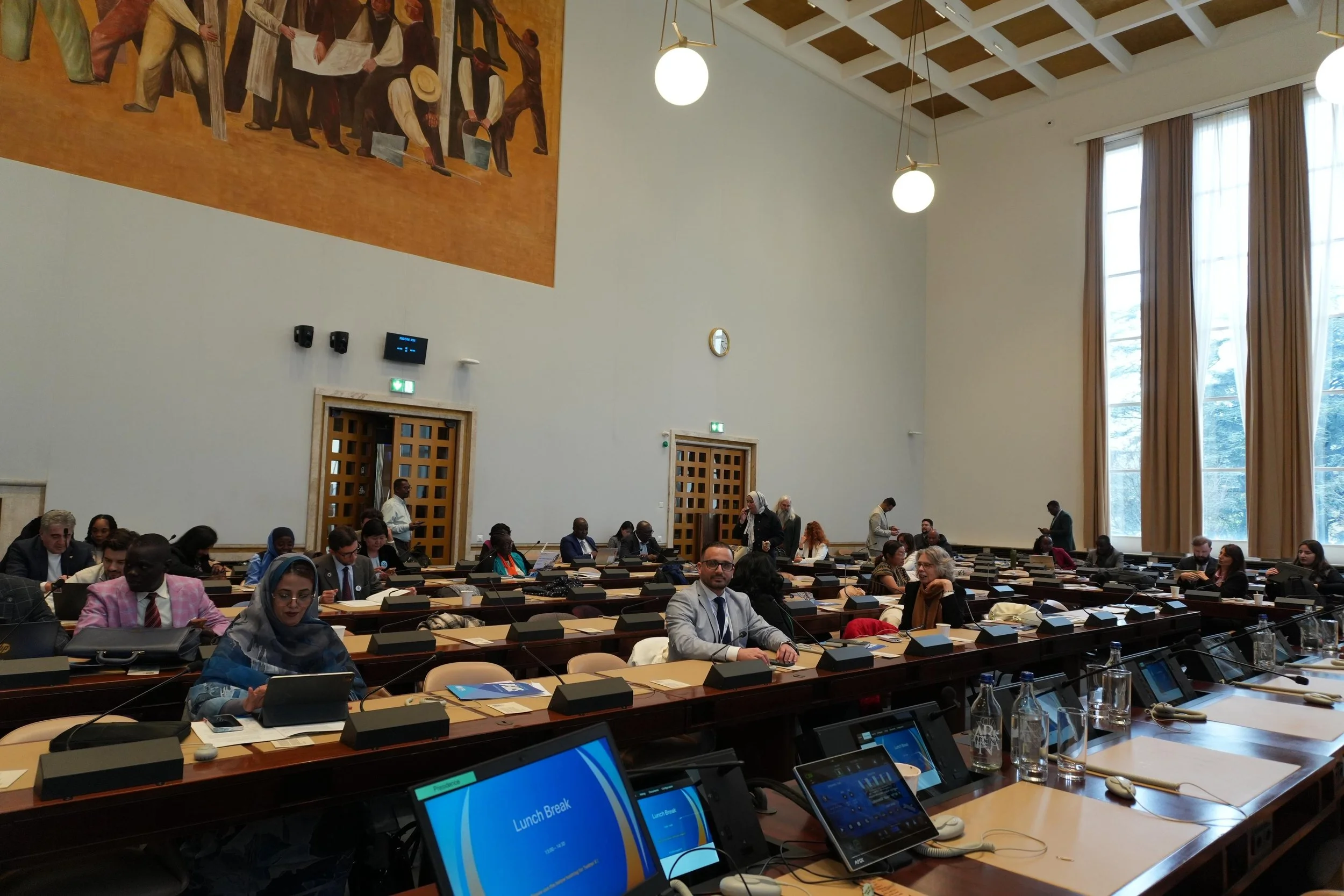 A large conference room filled with diverse people seated at long tables with microphones and laptops, large windows with curtains on the right, high ceiling with hanging lights, and a large mural on the wall depicting people holding tools and working together.