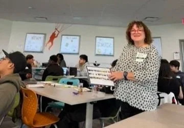 A woman standing in a classroom or training room holding a laptop, smiling, with students seated at tables working on computers or notebooks in the background, wall-mounted screens displaying presentations.