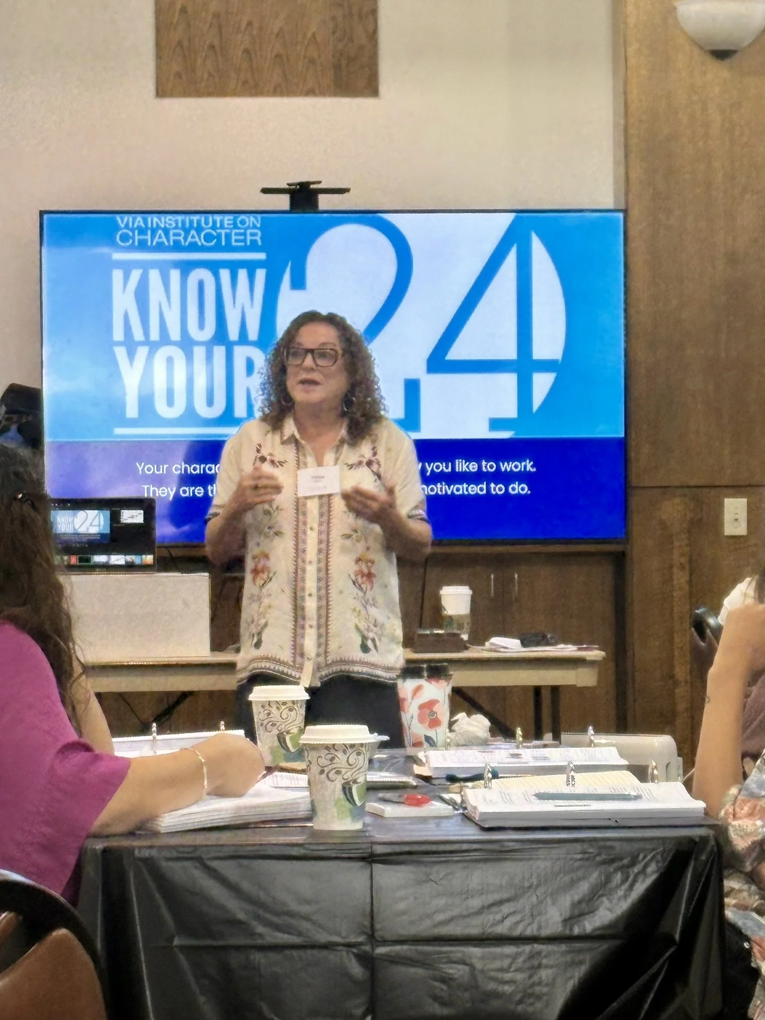 A woman with curly hair and glasses is giving a presentation at a workshop, standing in front of a screen that displays the text 'Know Your 24' and details about character development. Several people are seated at tables with notebooks, pens, and coffee cups.