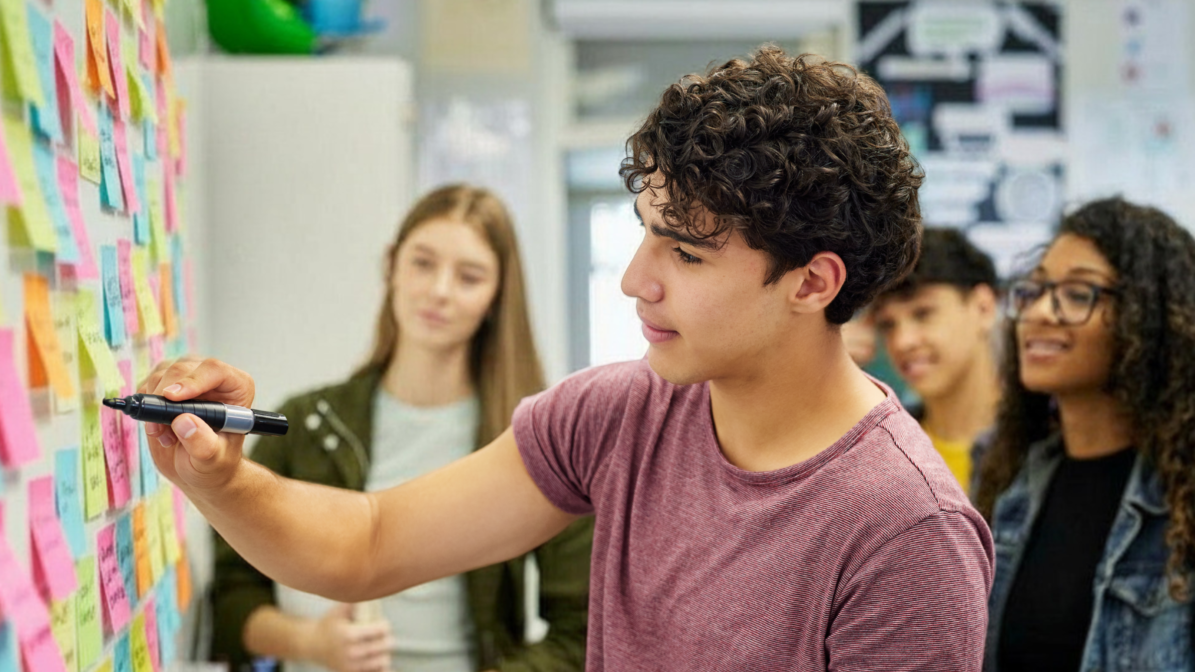 A young man with curly hair writing on a colorful sticky note wall while others observe, in an indoor setting.