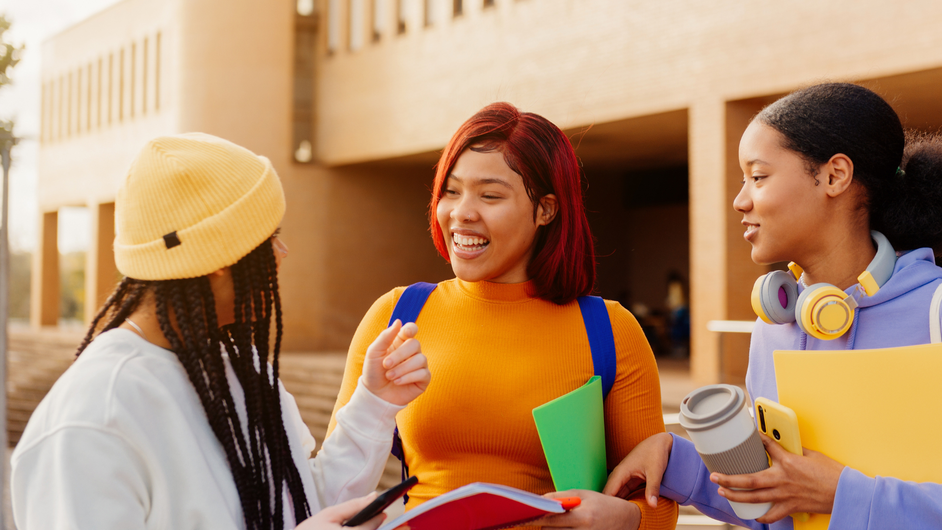 Three young women talking and smiling outdoors in front of a building, one with a yellow beanie, another with red hair, and the third with headphones around her neck, holding a coffee cup and stationery.