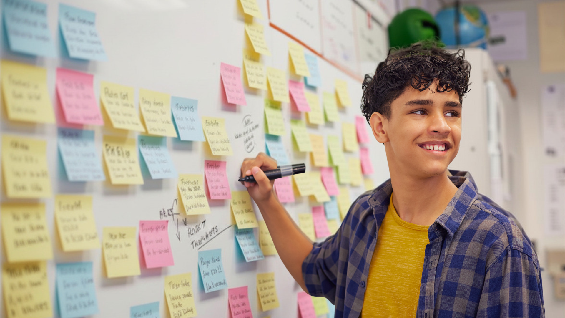 A young man with curly hair smiling and writing on a whiteboard covered with colorful sticky notes.