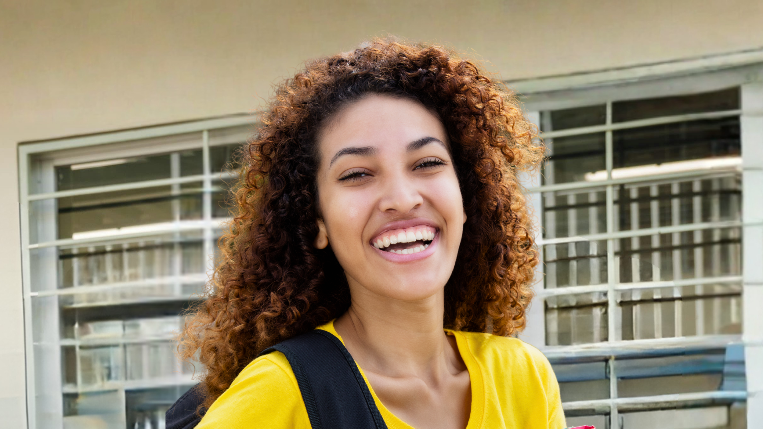 Close-up of a young woman with curly hair wearing a yellow shirt and backpack, smiling in front of a building with large windows.