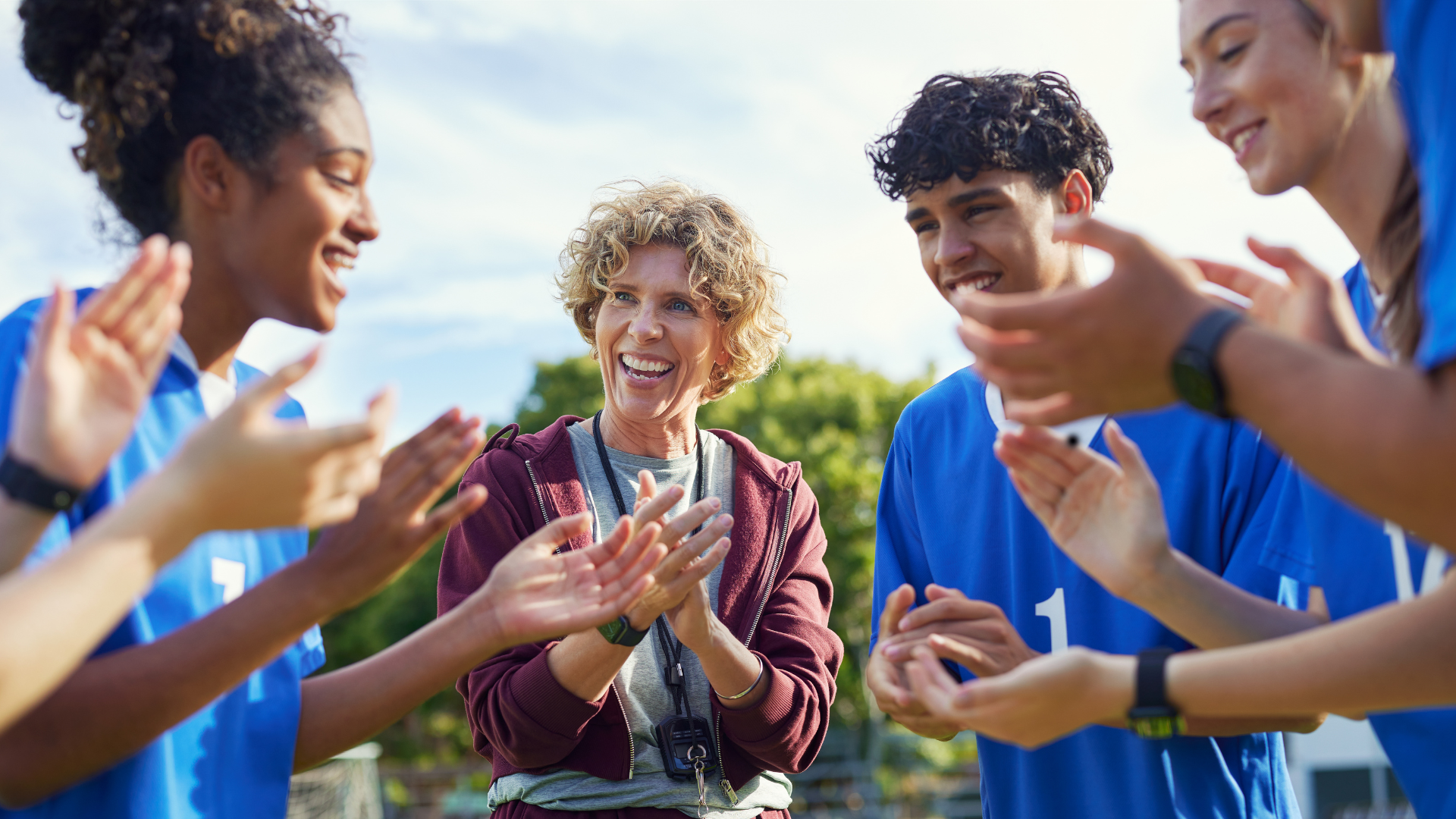 A coach with curly blonde hair smiling, surrounded by young female and male soccer players in blue jerseys, in an outdoor setting with trees and sky in the background.