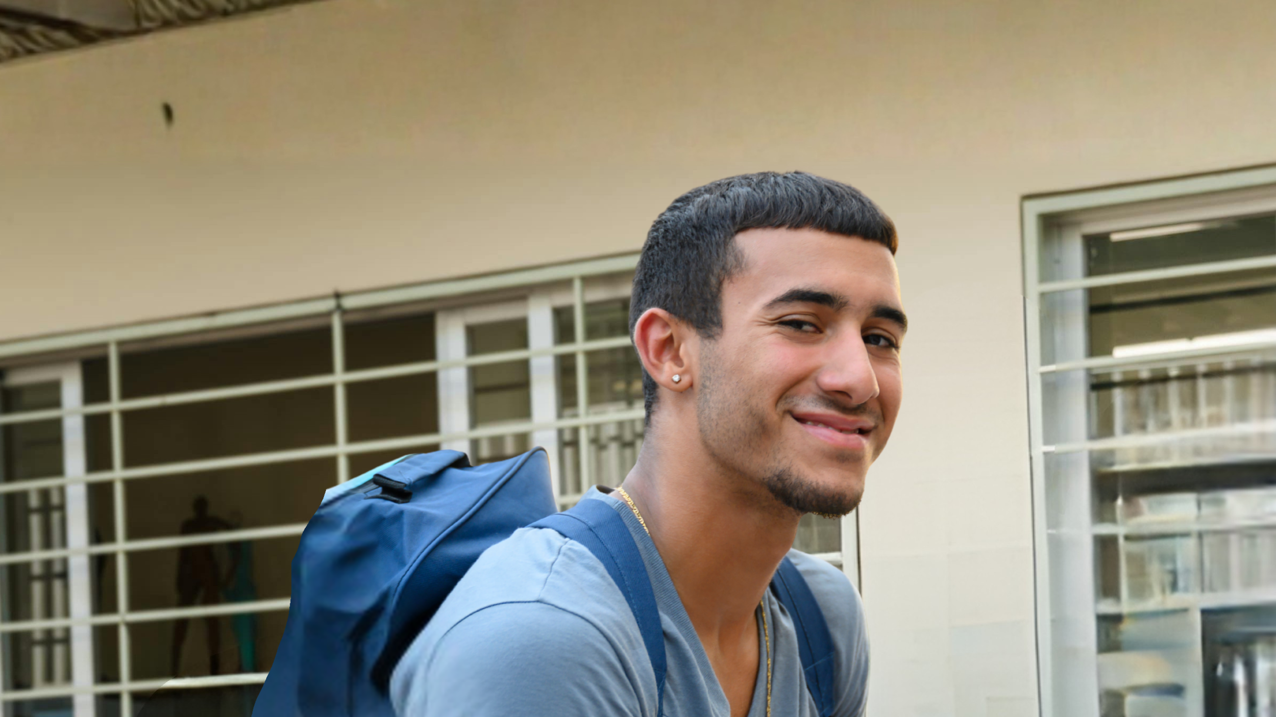A young man with short black hair, earrings, and a beard, smiling at the camera while carrying a blue backpack, standing outside a building with glass windows and white shutters.