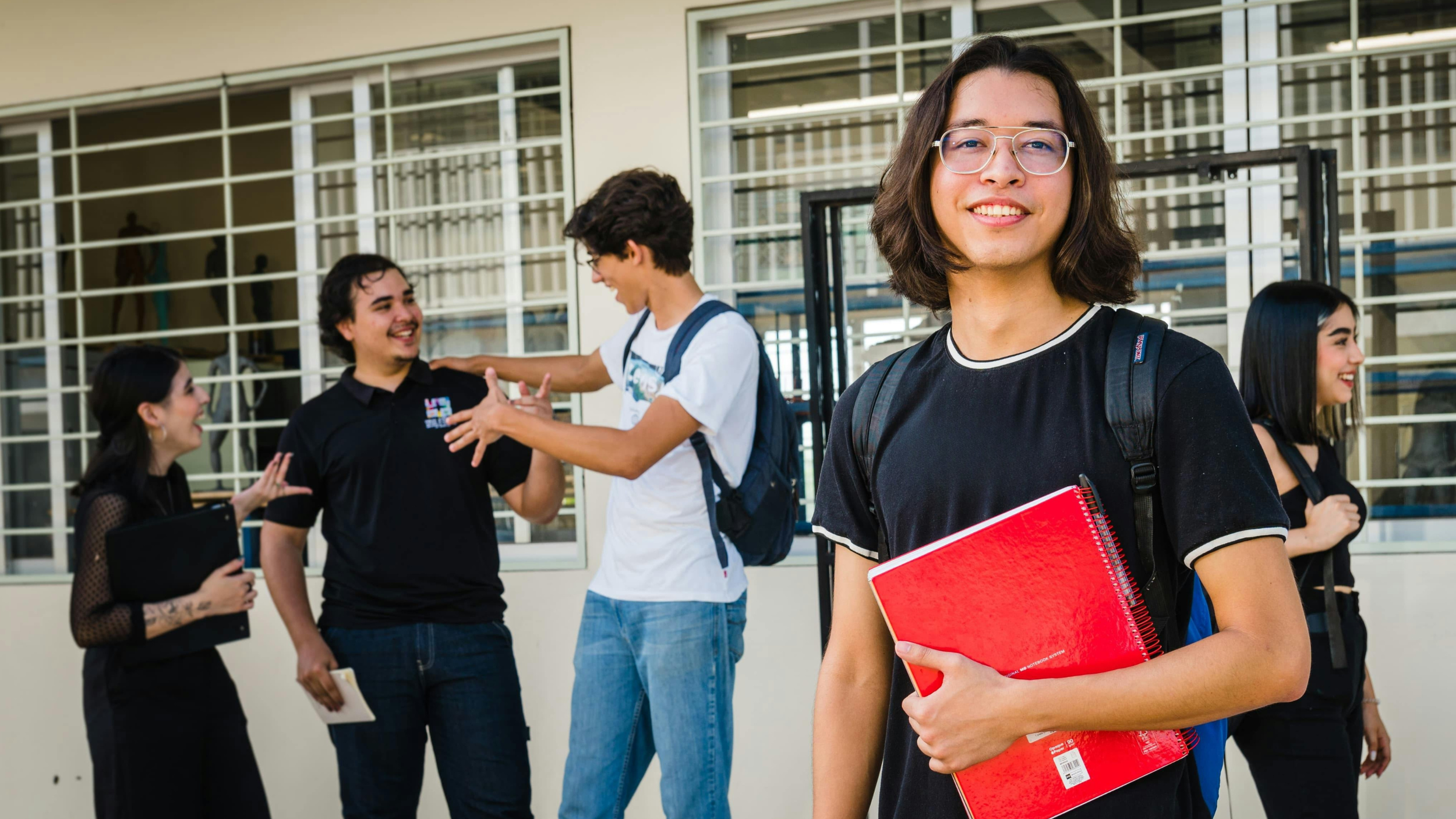 A group of five diverse students standing outside, engaging in conversations and smiling, with a modern building with large glass windows in the background. One student stands in front with a red notebook.