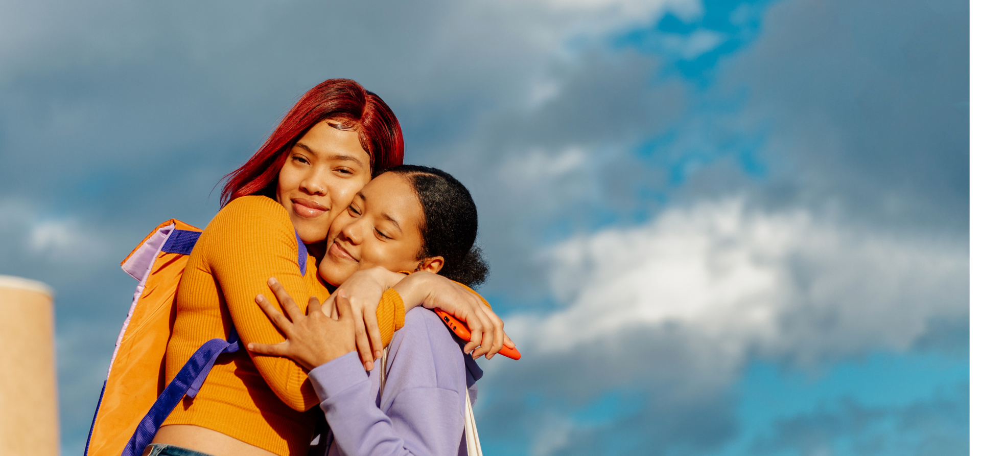 Two young women hugging and smiling outdoors under cloudy sky, one with red hair and a backpack, the other with black hair and a purple jacket.