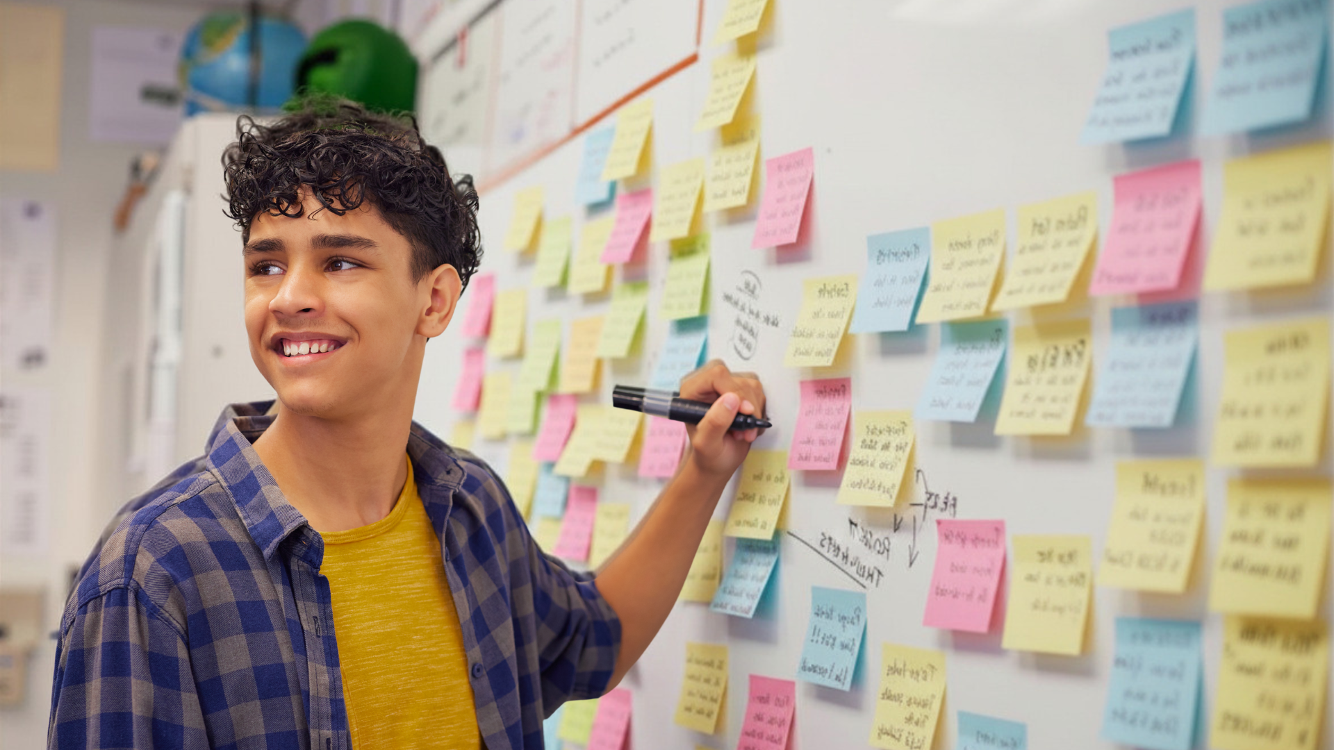 A young man with curly dark hair smiling as he writes on a whiteboard covered with colorful sticky notes.