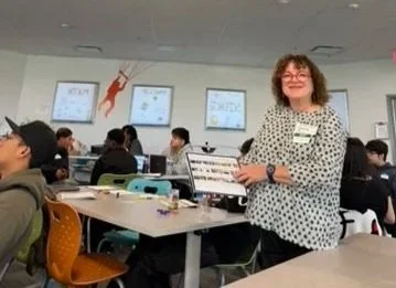 A woman standing in a classroom with students seated at desks, using laptops. The woman is smiling and holding a book, wearing glasses and a polka-dot shirt.