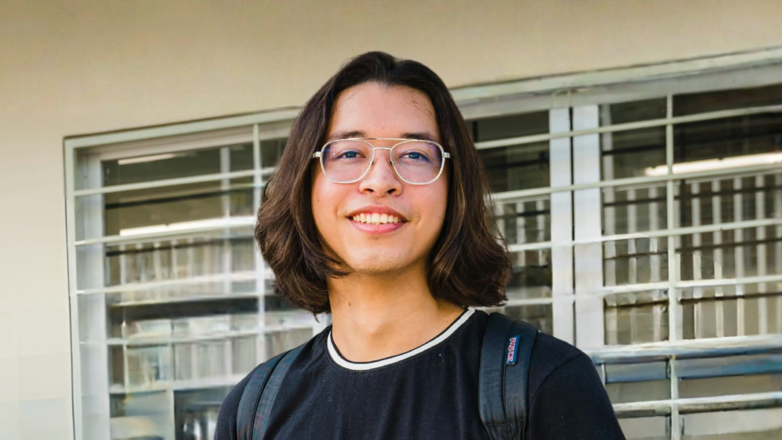 A young person with shoulder-length dark hair, glasses, and a black shirt, smiling outside in front of a modern building with horizontal window blinds.