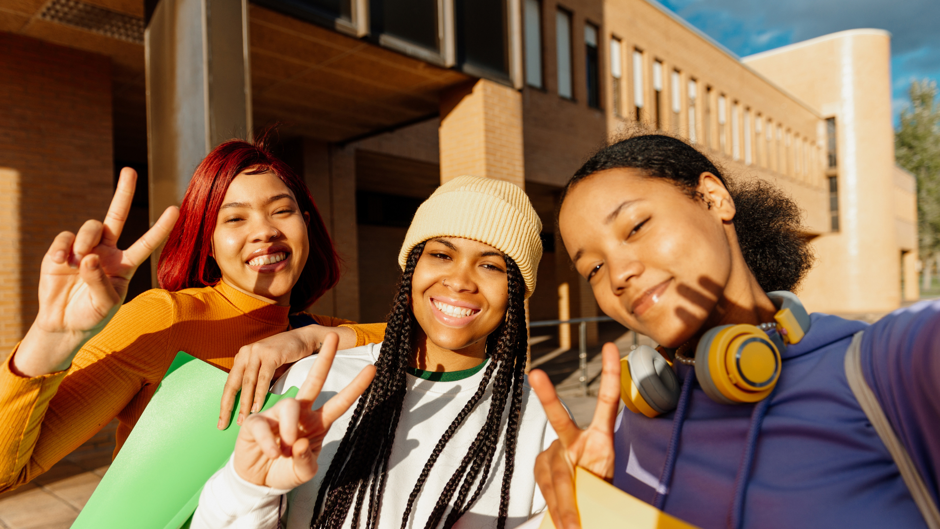 Three smiling girls taking a selfie outdoors, making peace signs, in front of a modern brick building on a sunny day.
