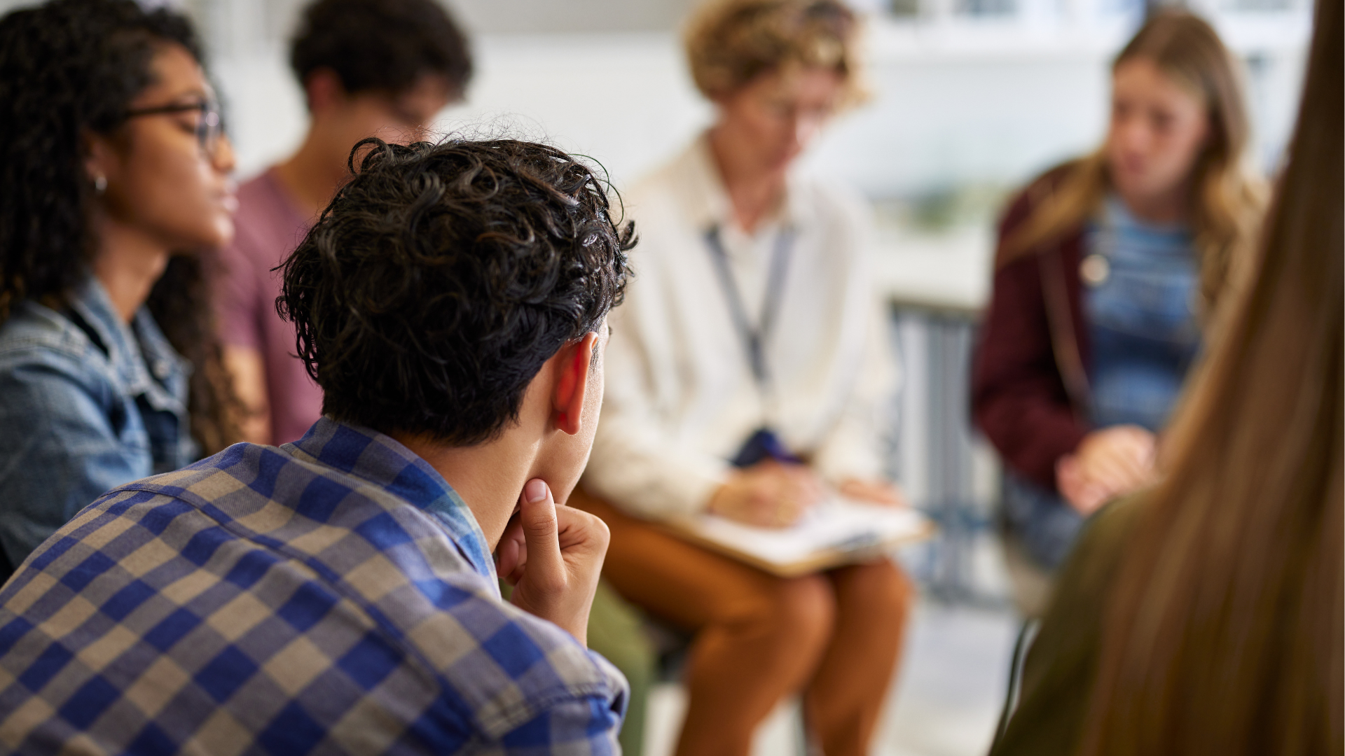 A group of young women and one man having a discussion in a bright room. The person in the foreground is a young man with dark, curly hair wearing a checkered shirt, resting his chin on his hand. The background shows three young women, one with blond hair, two with darker hair, all engaged in conversation, with one woman taking notes.