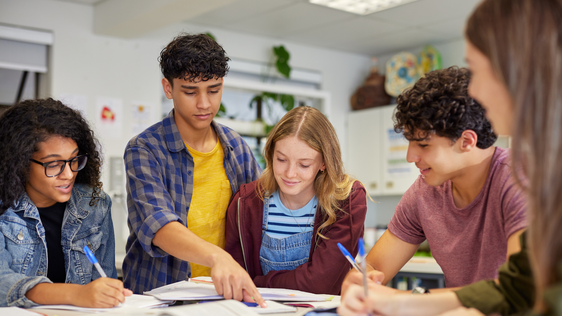Group of five teenagers studying and discussing at a table in a classroom or library, with books and notebooks open.