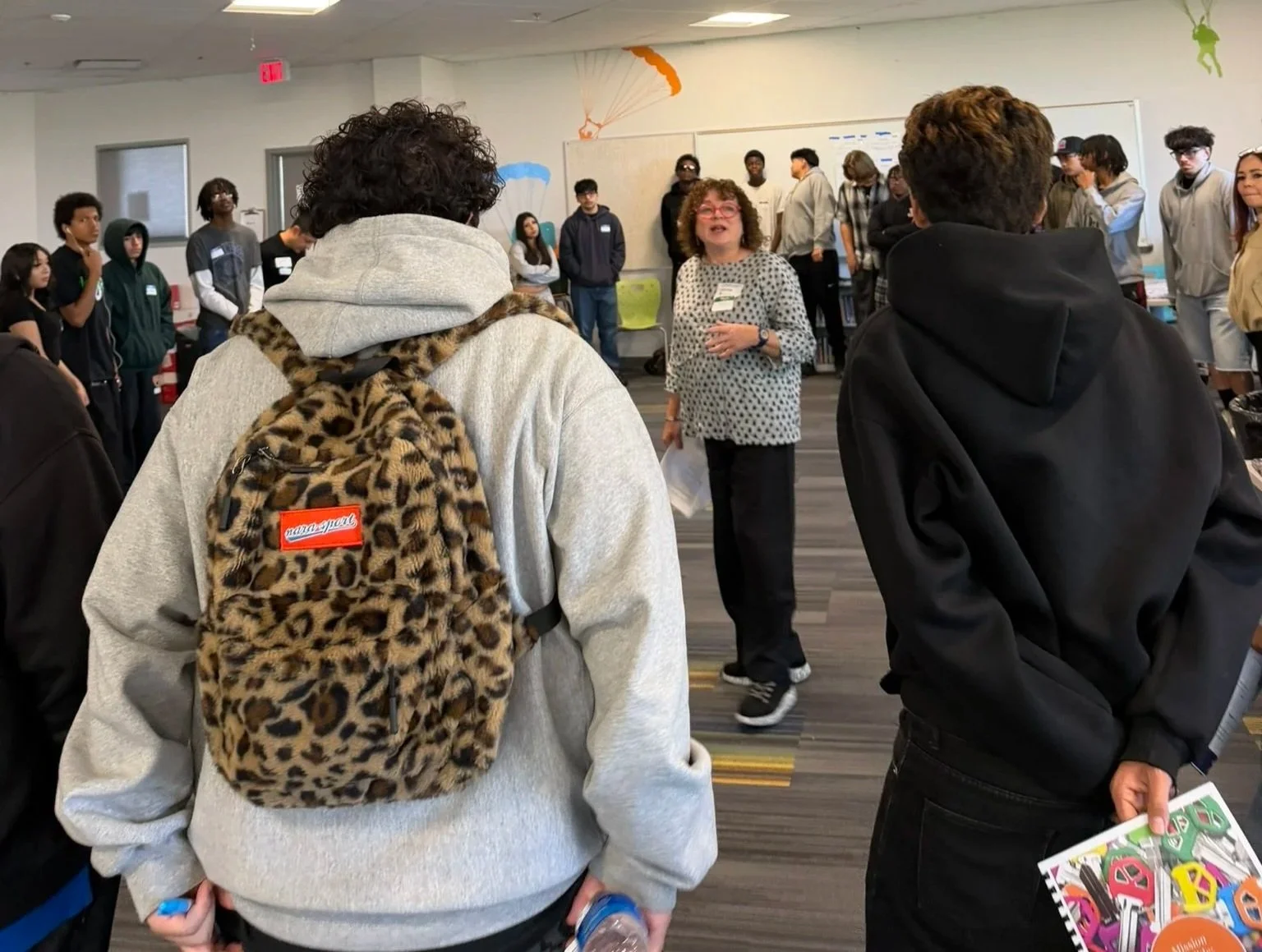 A group of diverse students listening to a woman speaking in a classroom or workshop.