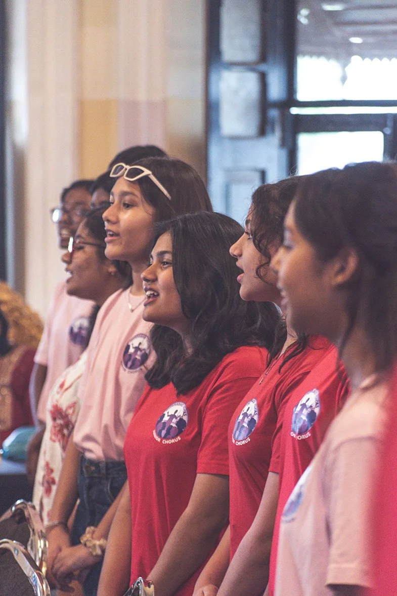 A group of women standing side by side, wearing matching shirts with a logo, possibly singing or participating in a choir event indoors.