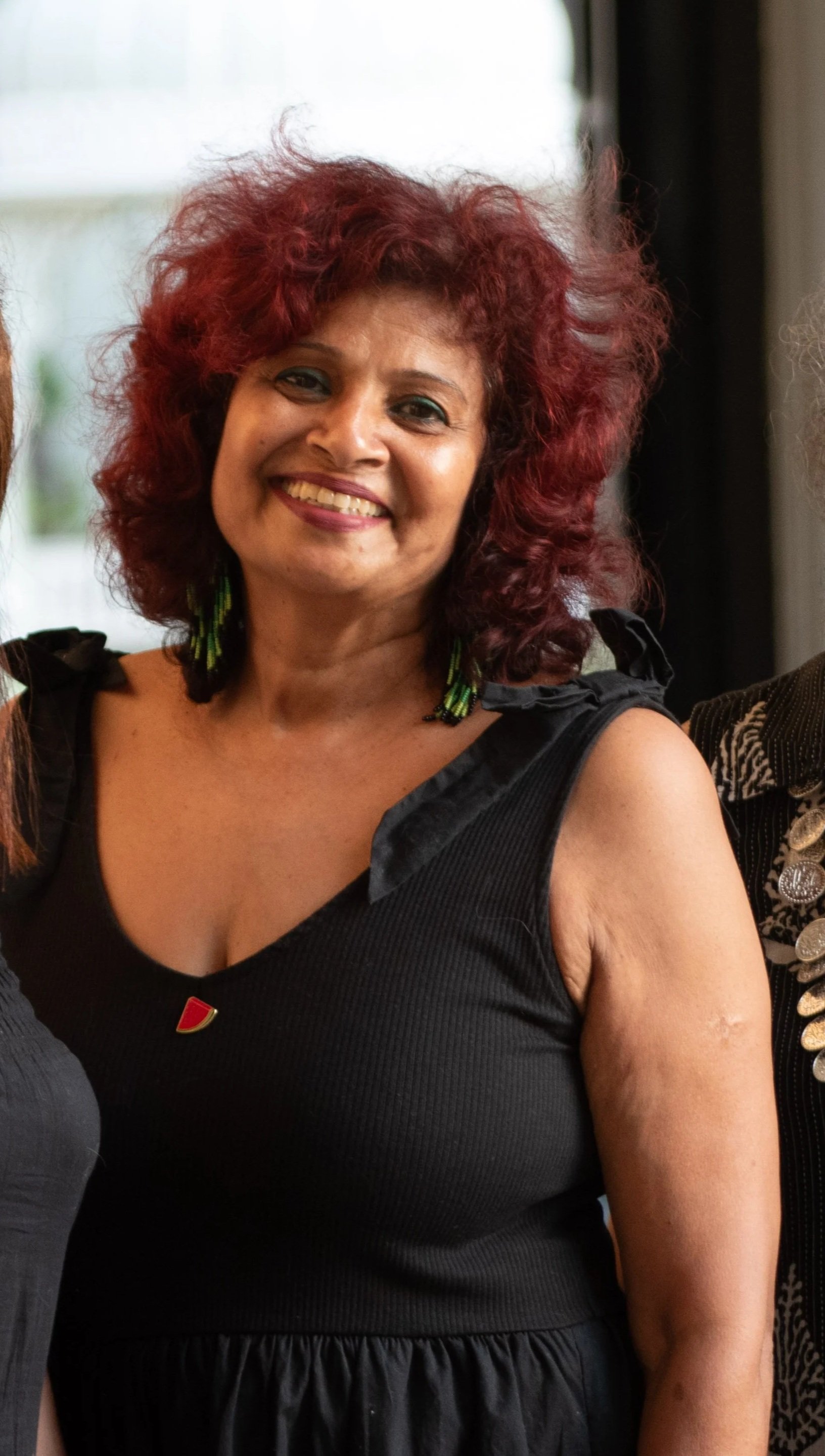 A smiling woman with curly red hair wearing a black sleeveless dress and green earrings.