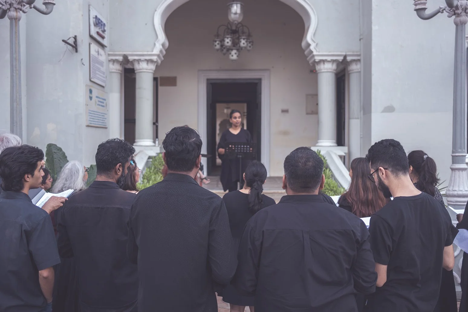 A group of people attending a gathering outside, with a woman speaking at a lectern in front of a historic building with white architectural features.