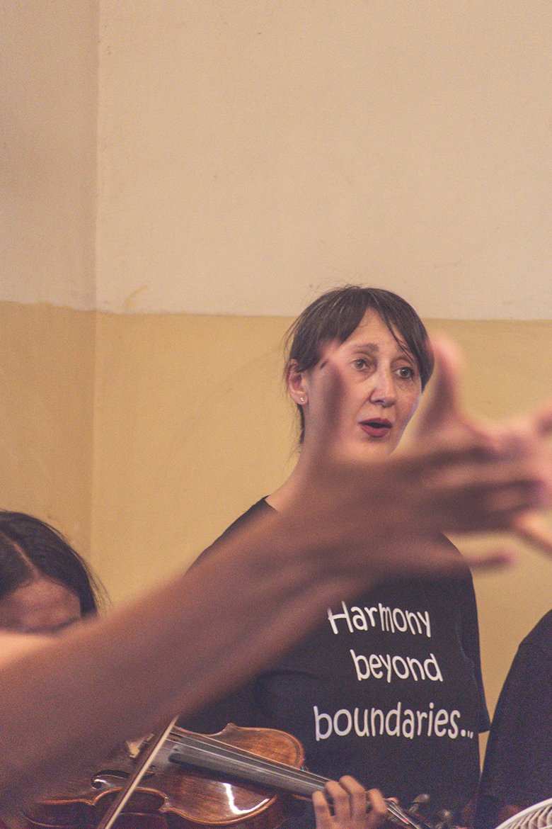 A woman with short dark hair singing, with a hand reaching out in front of her and holding a violin, in a room with beige walls. She is wearing a black shirt with the words "Harmony beyond boundaries" written on it.