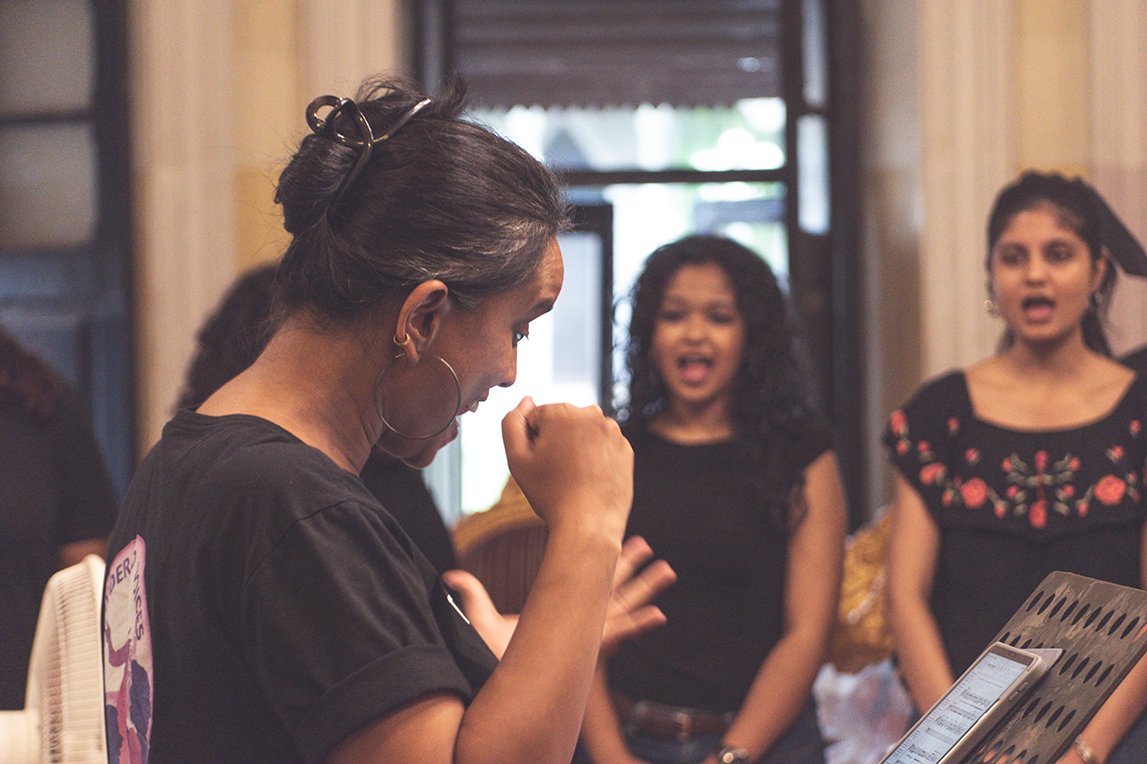 A woman with dark hair and hoop earrings is looking down and clenching her fist, while three other women in the background are singing passionately.