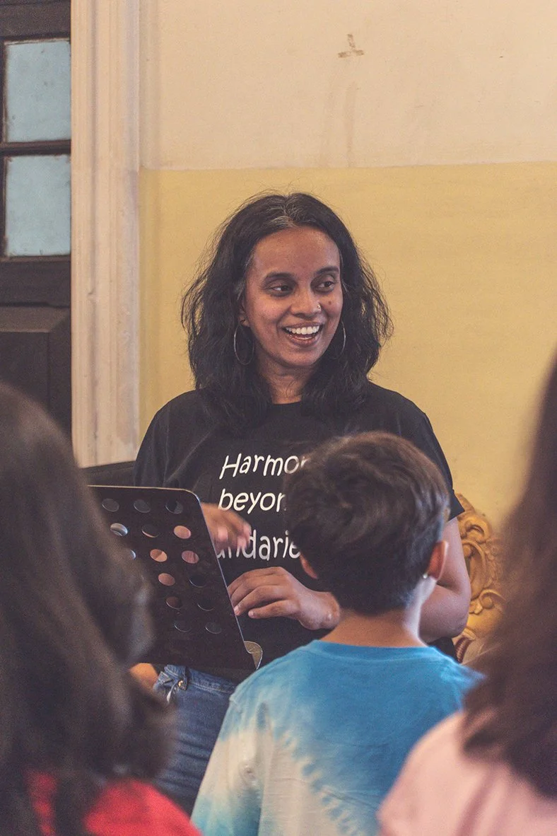 A woman with dark, curly hair smiling and wearing a black t-shirt with white text, surrounded by children, inside a room with beige and yellow walls and a window with dark wood framing.