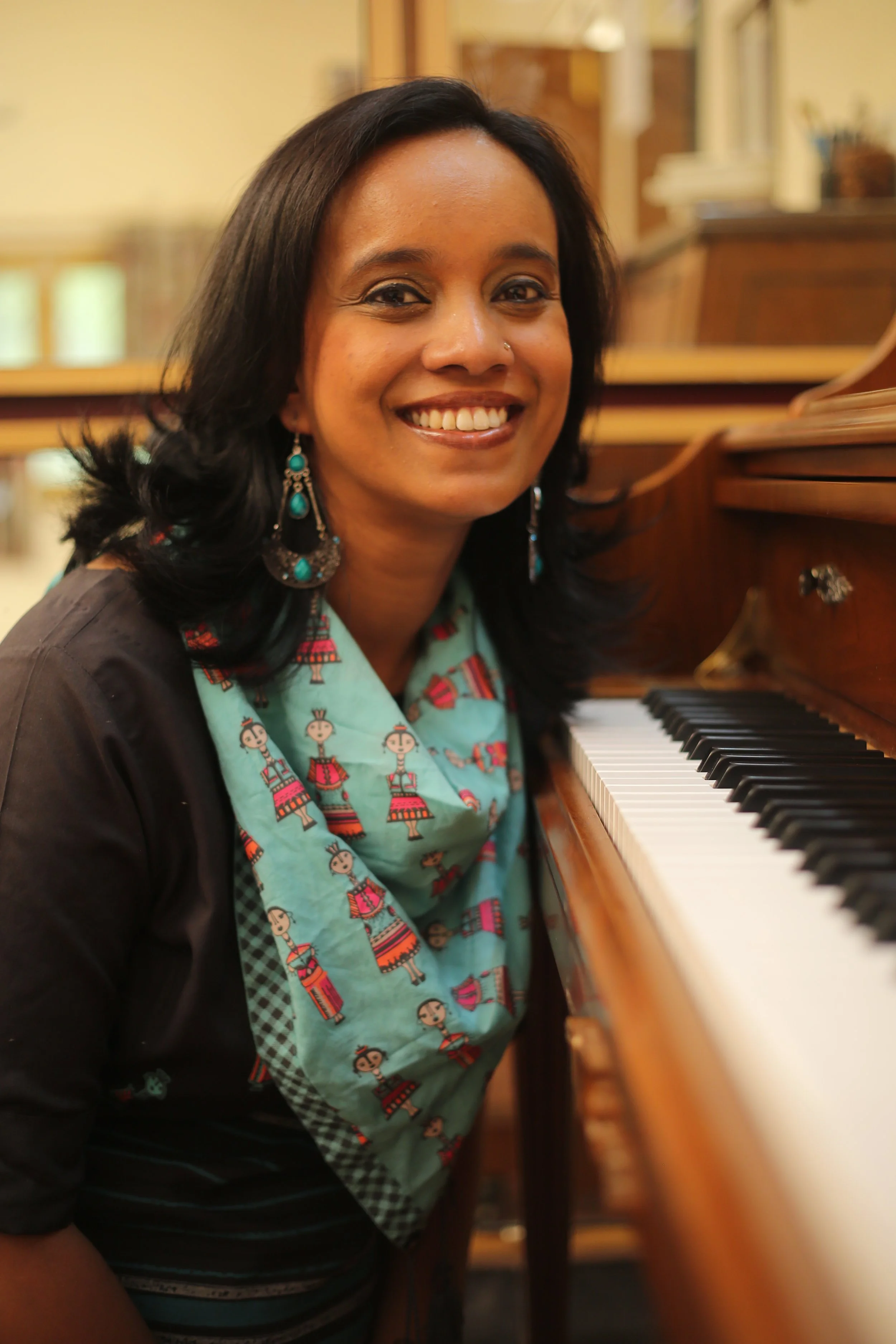 A smiling woman with black hair and earrings, sitting at a wooden piano, wearing a teal shirt with a pattern of dancers, in a cozy room with yellow walls and wooden furniture.