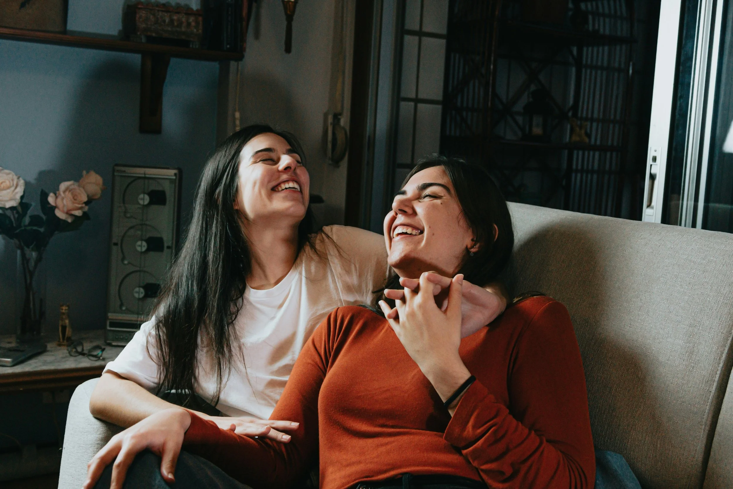 Two young women with long dark hair laughing and hugging on a couch in a living room.