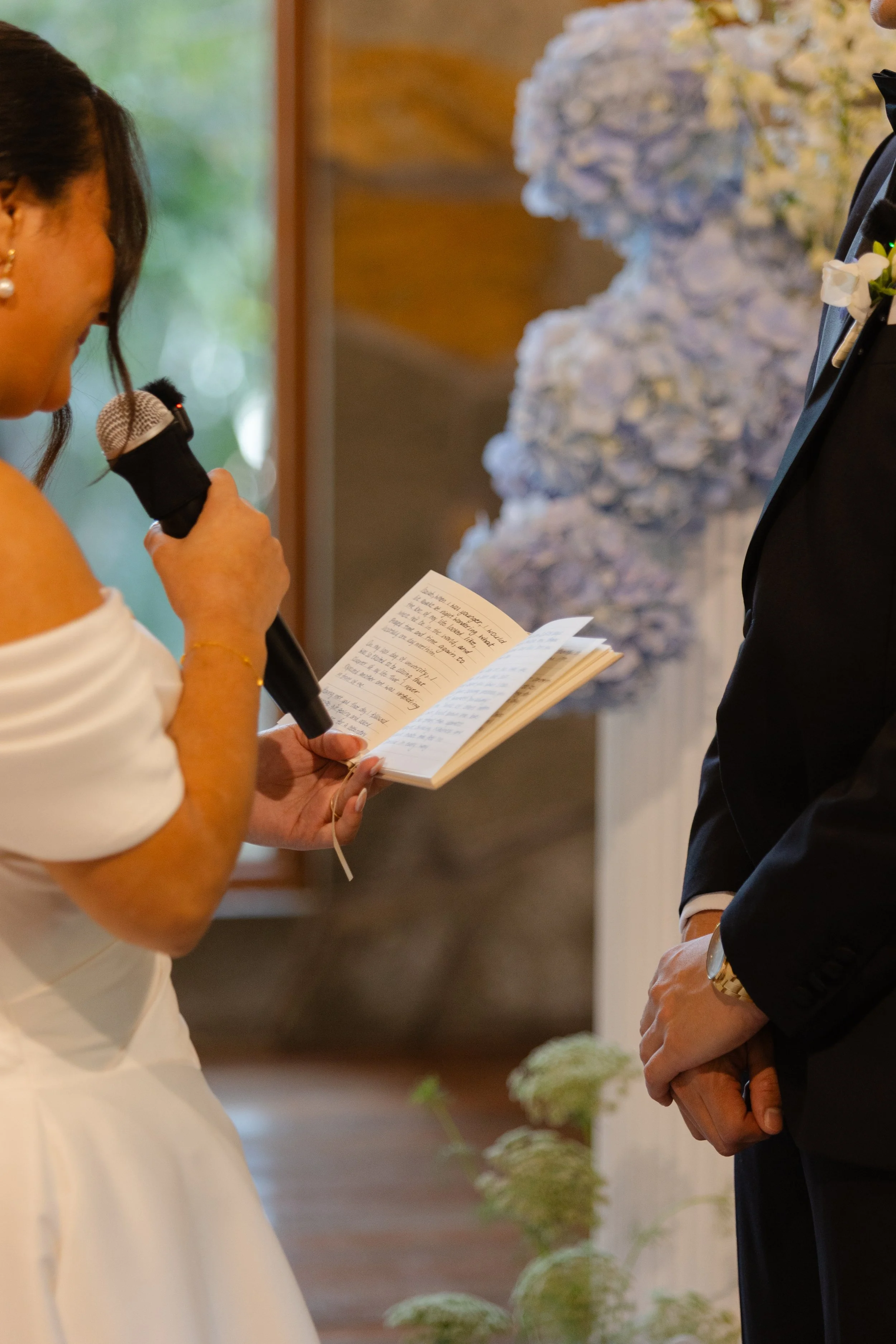 A woman in a white dress is reading from a book while holding a microphone, standing in front of a man in a suit during a wedding ceremony. There are white flowers in the background.