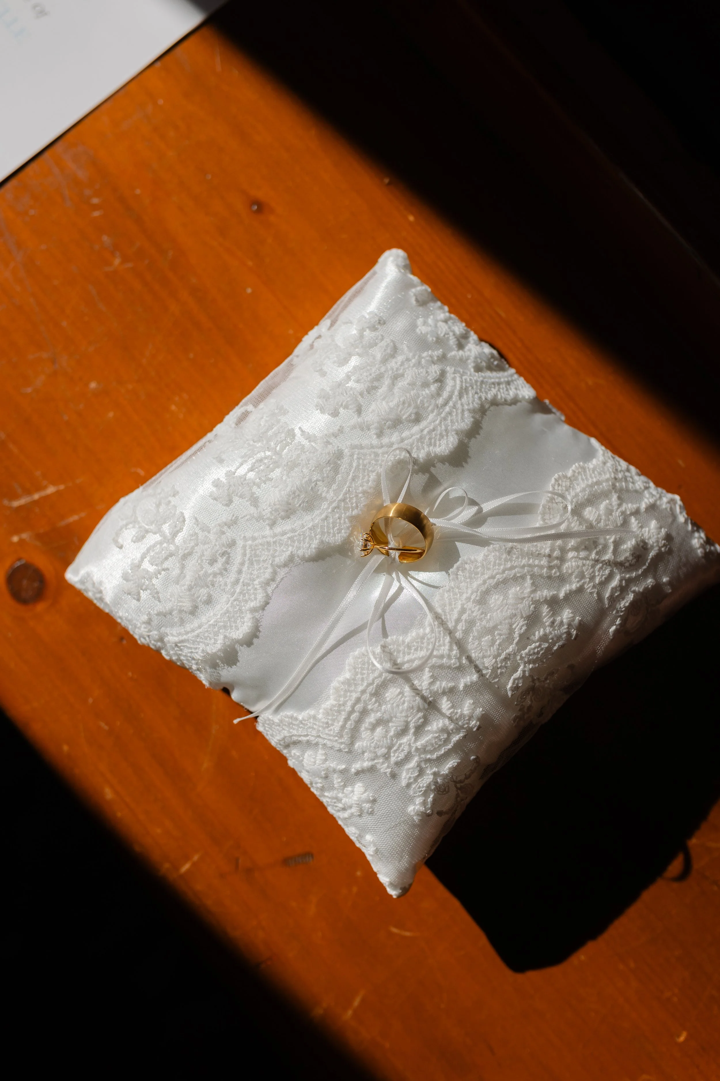 A white lace pillow with a wedding ring on top, decorated with a satin ribbon bow and floral embroidery, placed on a polished wooden surface.
