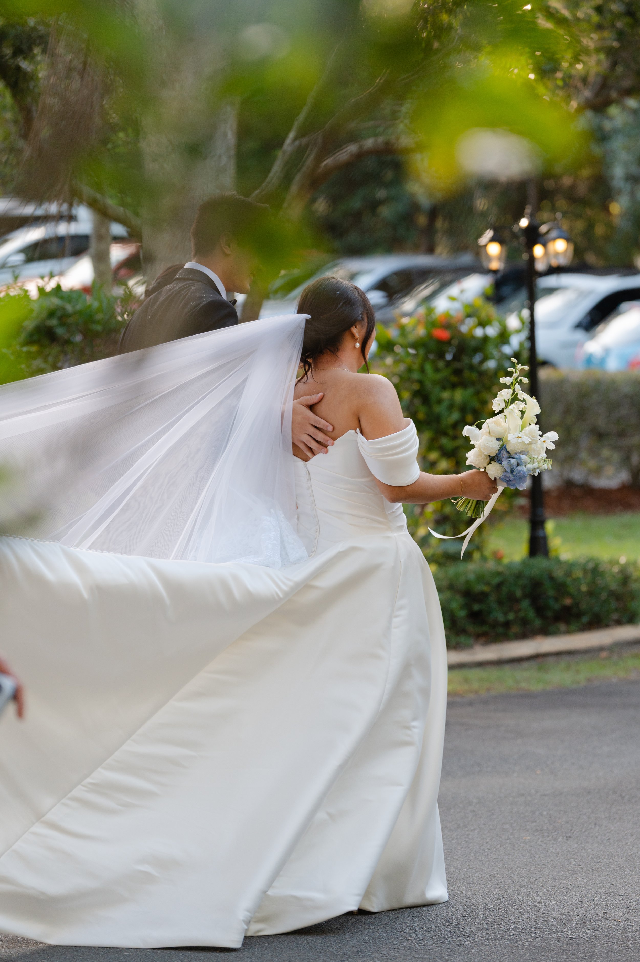 A bride in a wedding gown holding a bouquet standing outdoors with a groom in a tuxedo behind her, partially obscured by leaves and a wedding veil flowing behind her.
