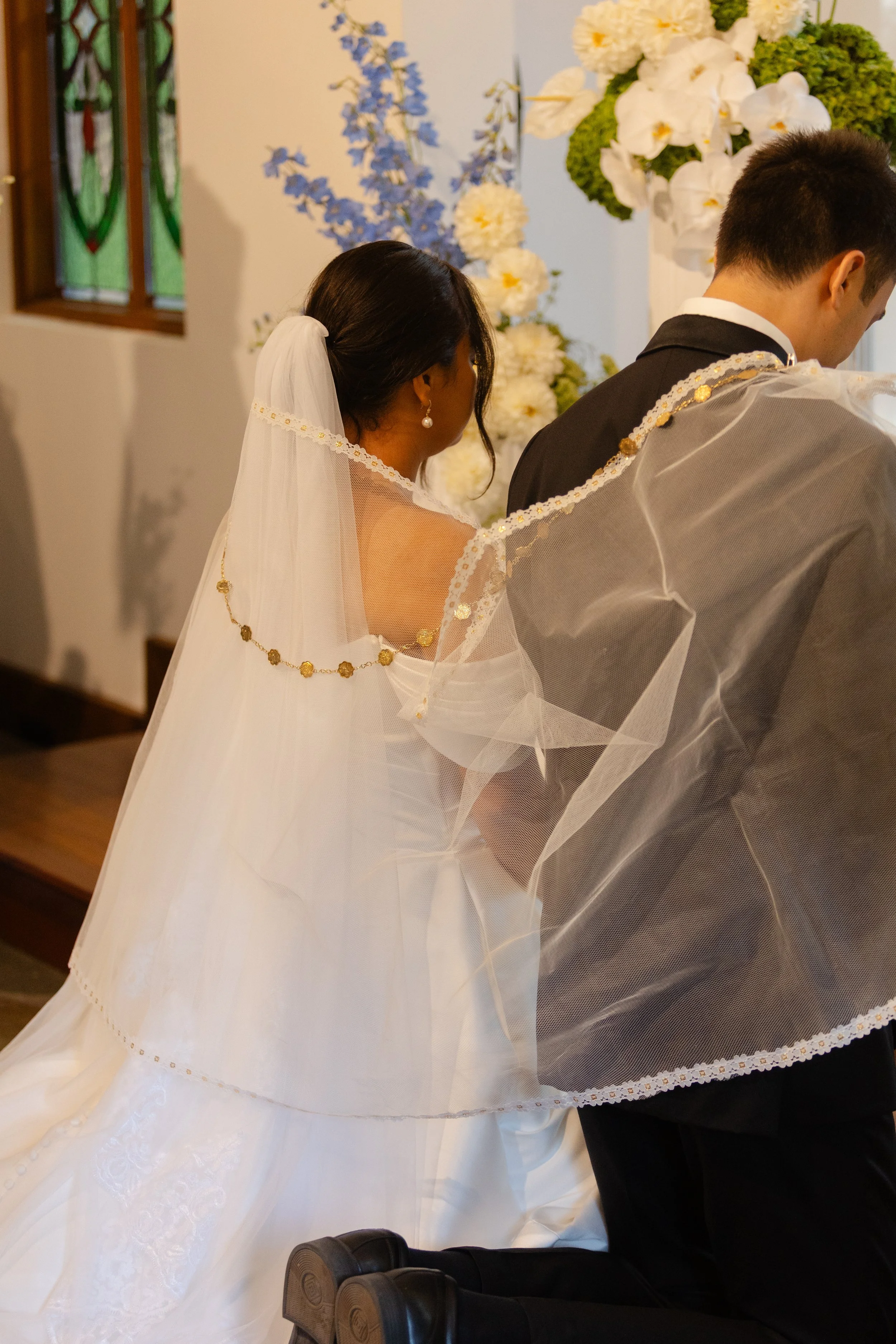 A bride and groom kneeling during a wedding ceremony inside a church, with floral arrangements and stained glass windows in the background.