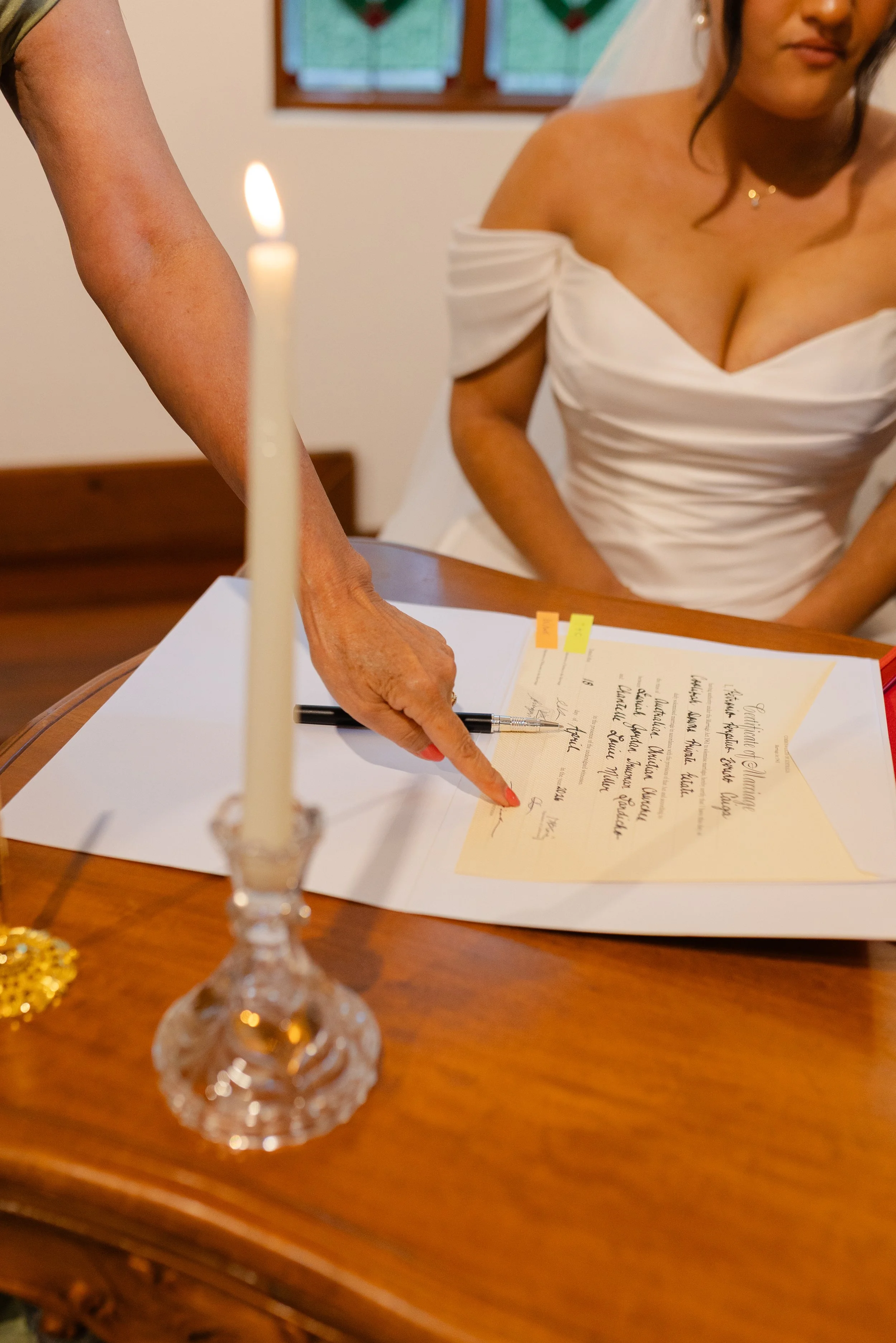 A bride in a white off-shoulder wedding dress signing a document with a black pen, with a lit candle and a decorated table in the foreground.