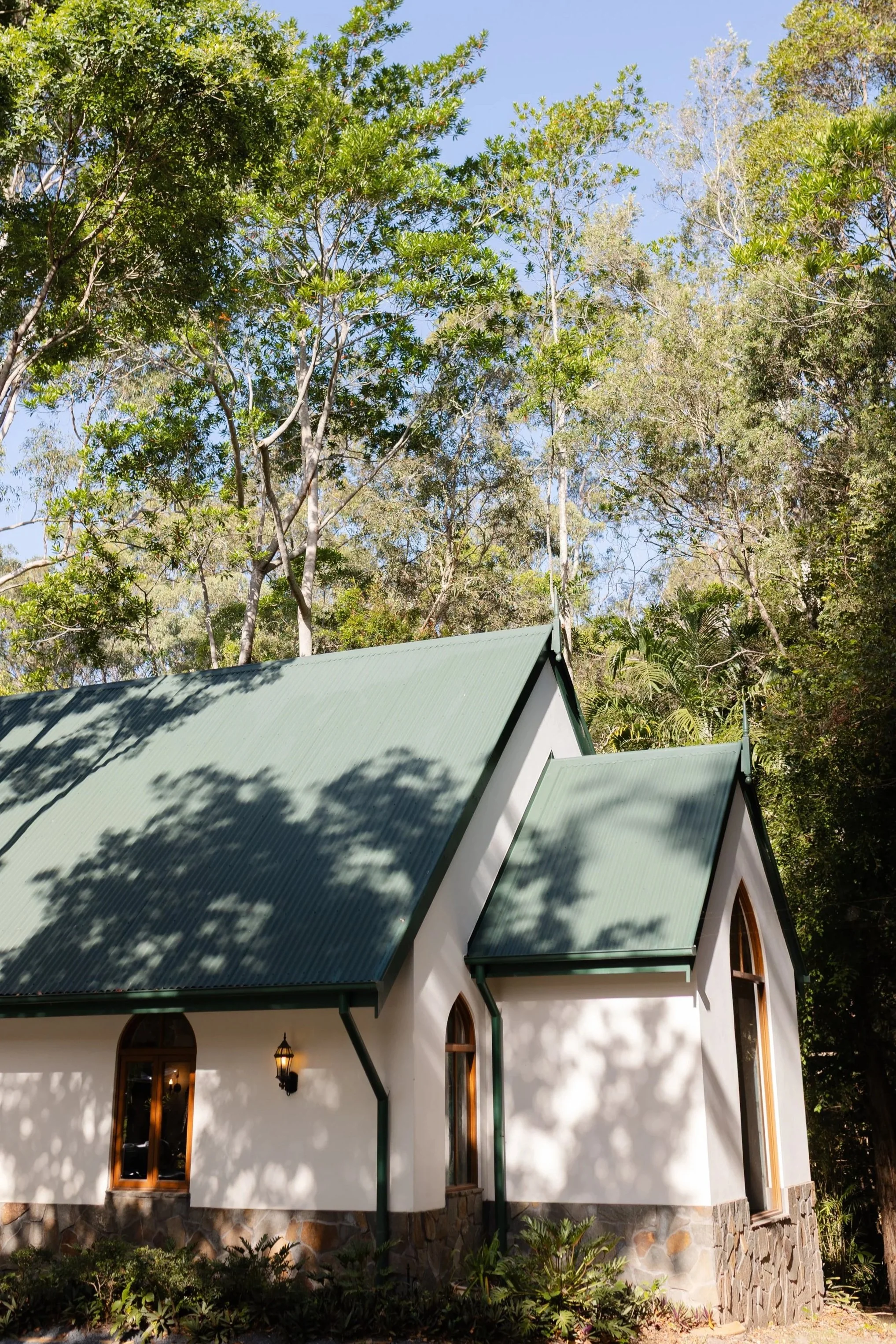 A white house with a green metal roof, arched windows, and exterior lights, surrounded by tall trees and foliage under a clear blue sky.