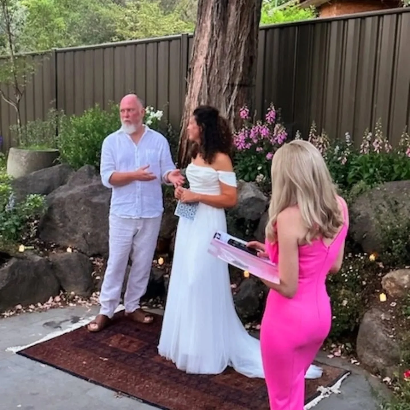 A wedding ceremony outdoors with a man and woman standing on a rug, facing each other, with a woman in a pink dress holding a clipboard nearby. The bride is in an off-shoulder white dress, and the groom wears a white shirt and pants. There are plants, flowers, and rocks in the background, with a large tree behind the couple.