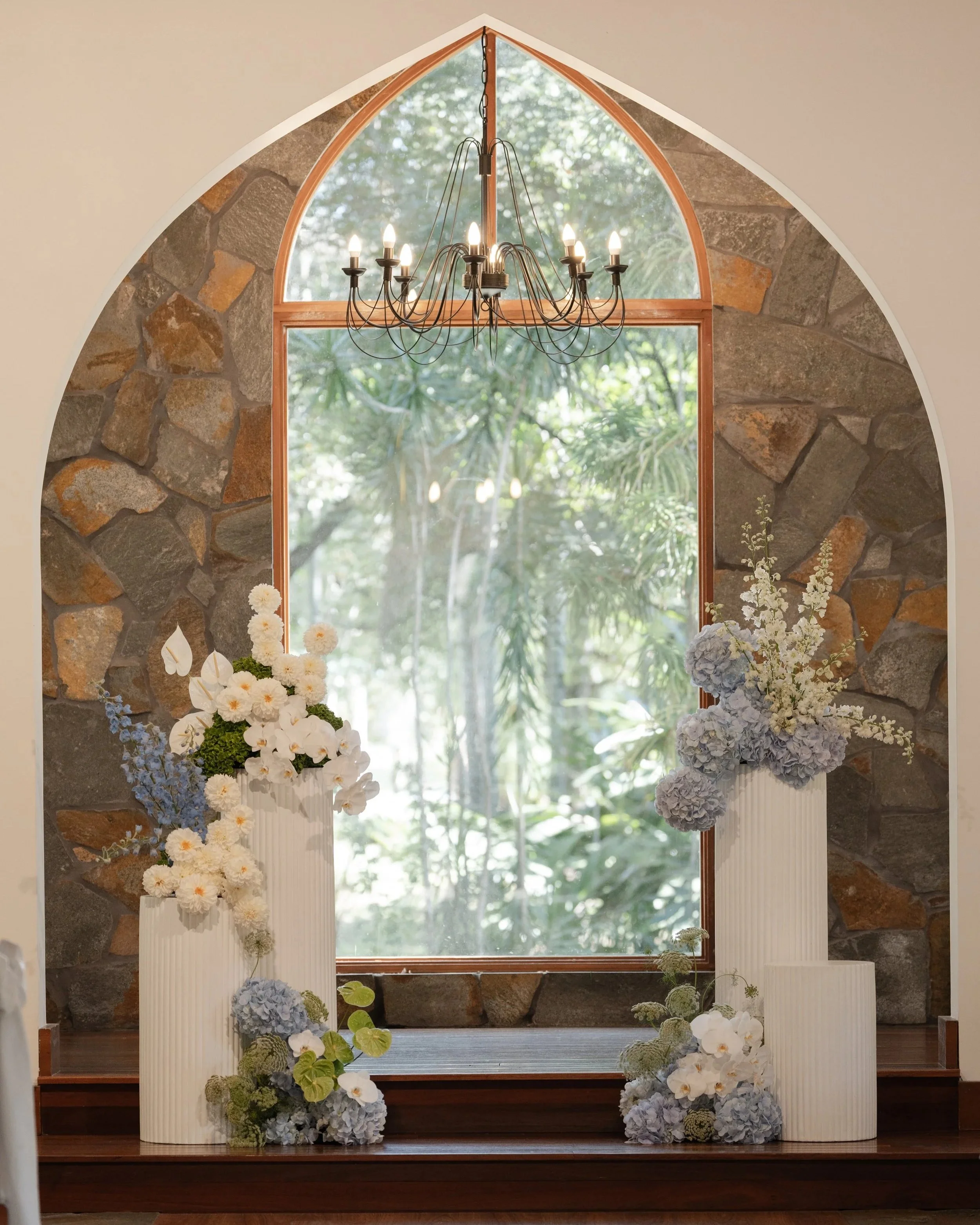 Chandelier hanging in front of a large window with stone walls on either side, decorated with white and light blue hydrangeas and other flowers.