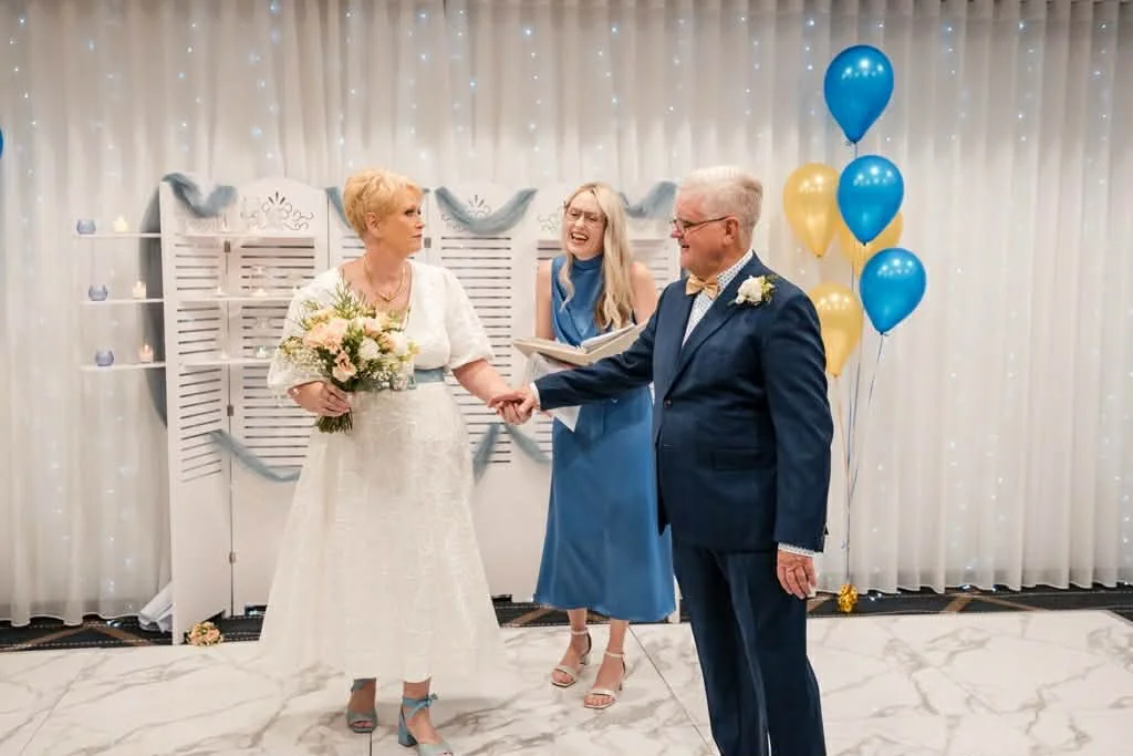An elderly couple exchanging vows or rings at a wedding or renewal ceremony, with a woman officiant standing behind them, in a decorated venue with balloons and twinkling lights.
