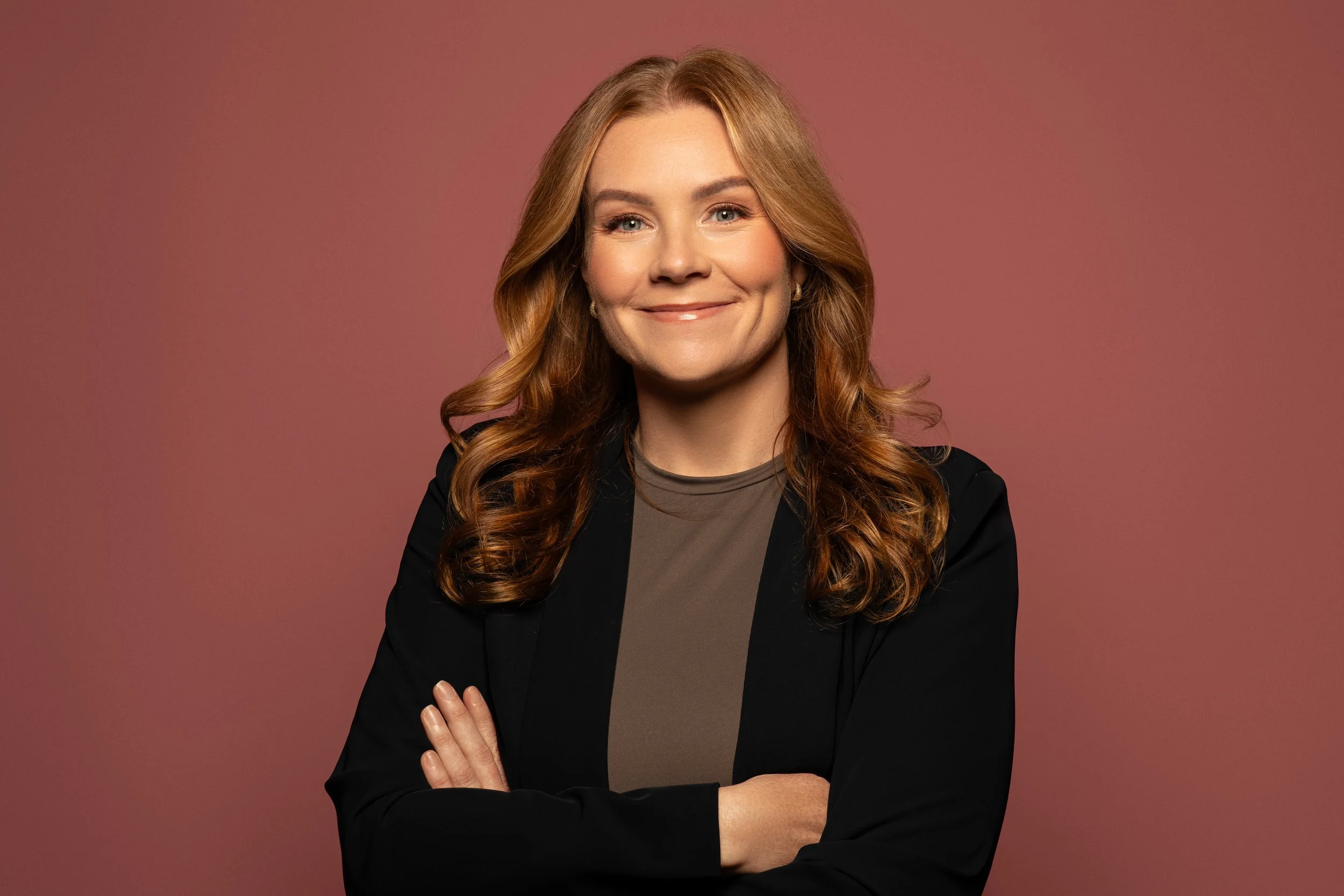 Portret van een vrouw met lang golvend rood haar, glimlachend, gekleed in een zwarte blazer en een bruine top, staand met één arm over de borst tegen een roze achtergrond.