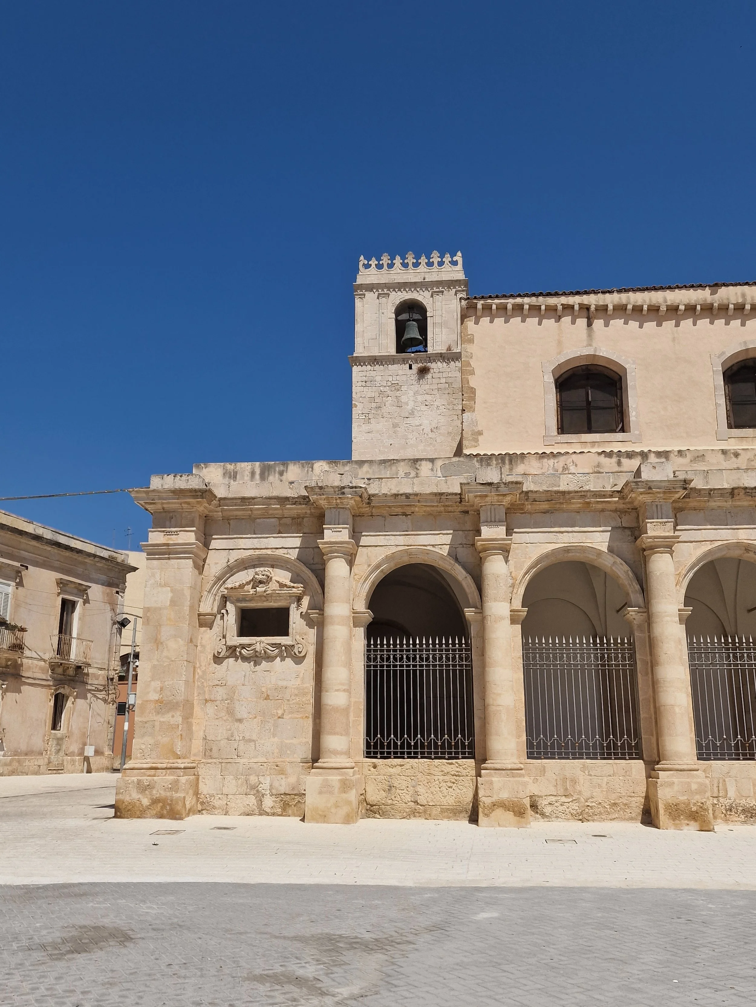 Sicily Ortigia Historic Center - Facade of an old stone building with arched windows, columns, and a bell tower with a bell and decorative crenellations against a clear blue sky.
