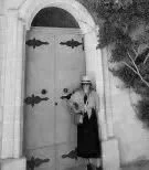 Selectedly Travel curator and local art-history specialised guide of Malta and Gozo Michelle Amato from Malta  in front of a large arched wooden door, holding a small bag, with trees nearby.