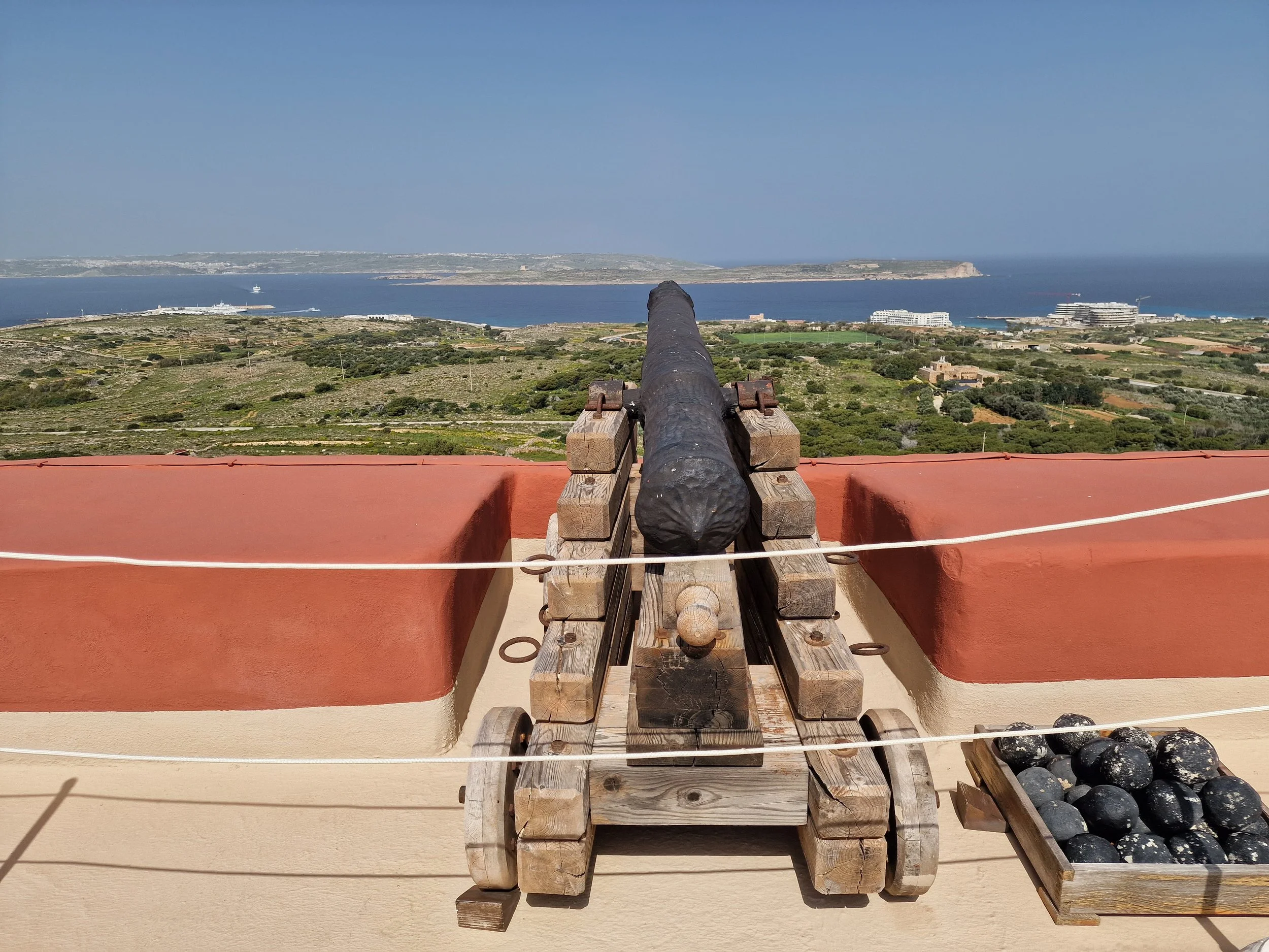 View of a coastal landscape with a cannon in the foreground and wooden sleds filled with cannonballs to the right.