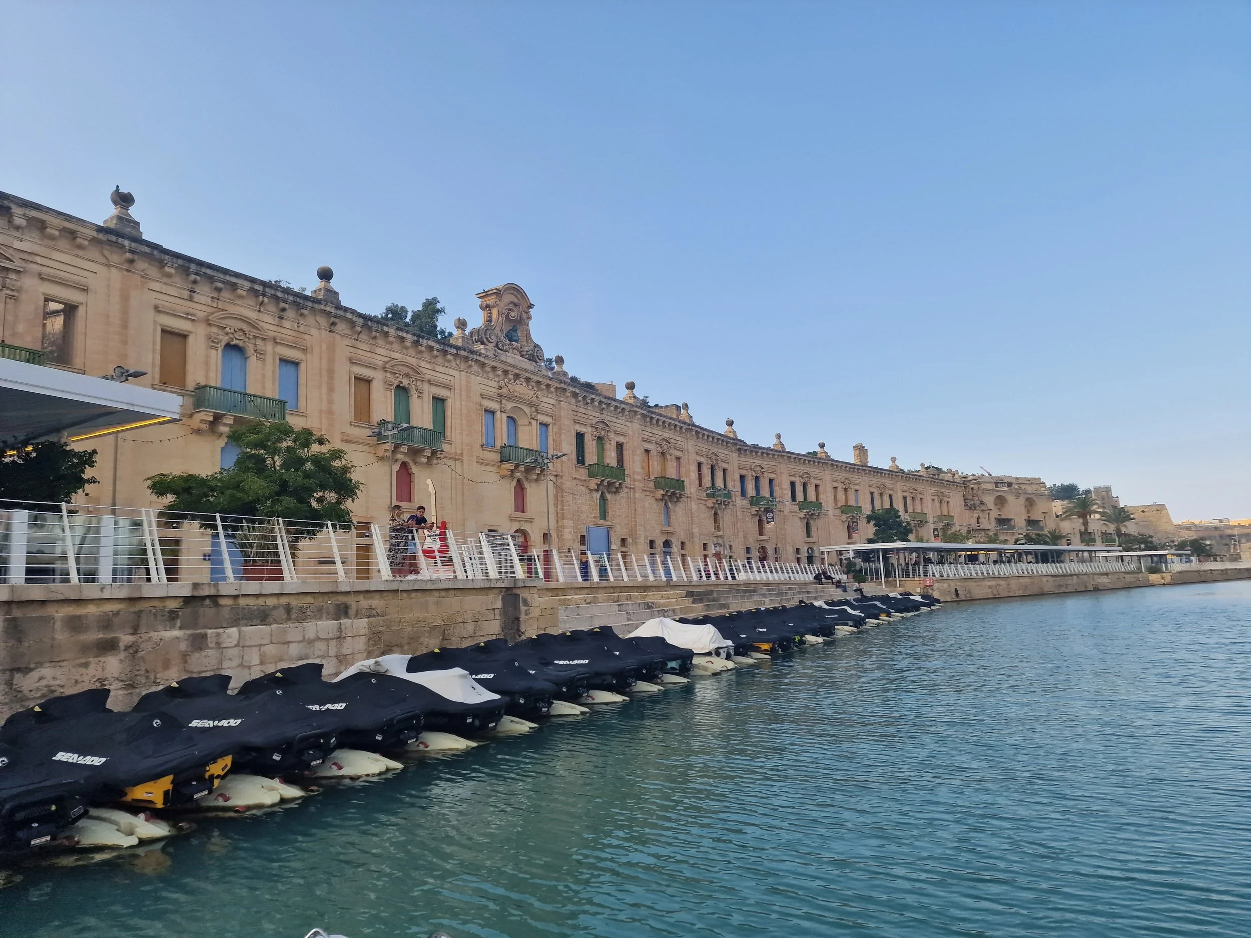Valletta Cruise-Liner Terminal - View of the historic buildings on Pinto Wharf Valletta Malta, along the waterfront with a row of jet skis parked in the water and a person walking along the promenade. This area used to be used by merchants.